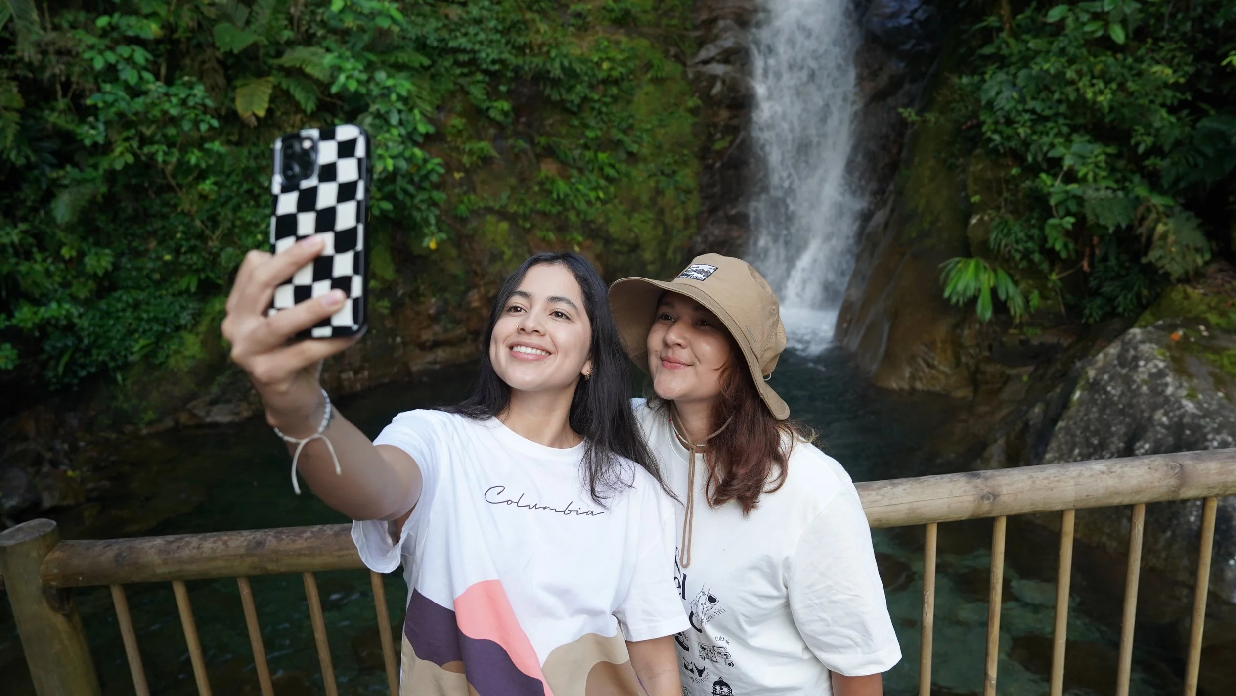 Dos mujeres toman una selfie junto a una cascada rodeada de vegetación.
