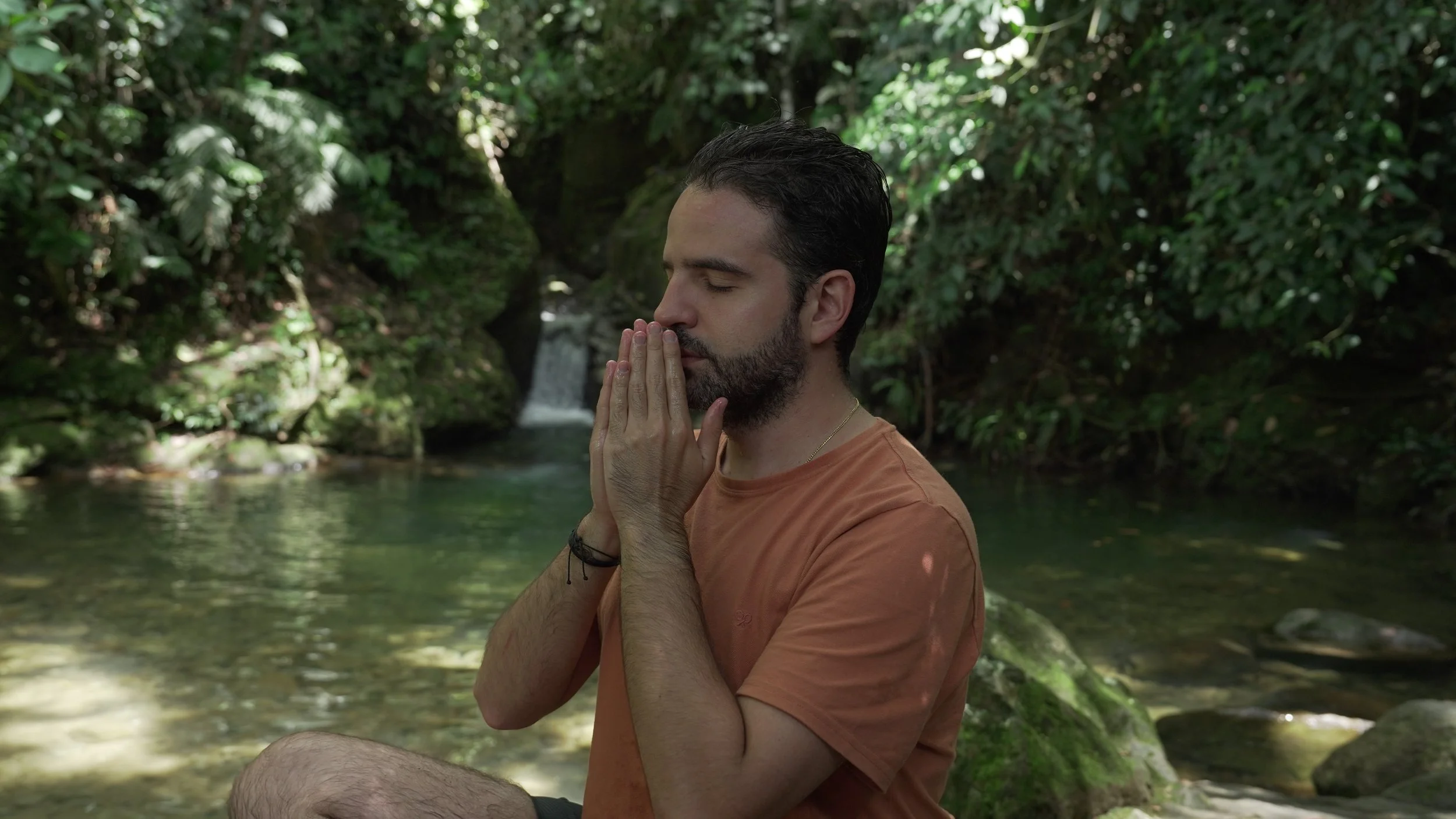 Hombre con barba en posición de meditación en un entorno natural junto a un río y vegetación densa.