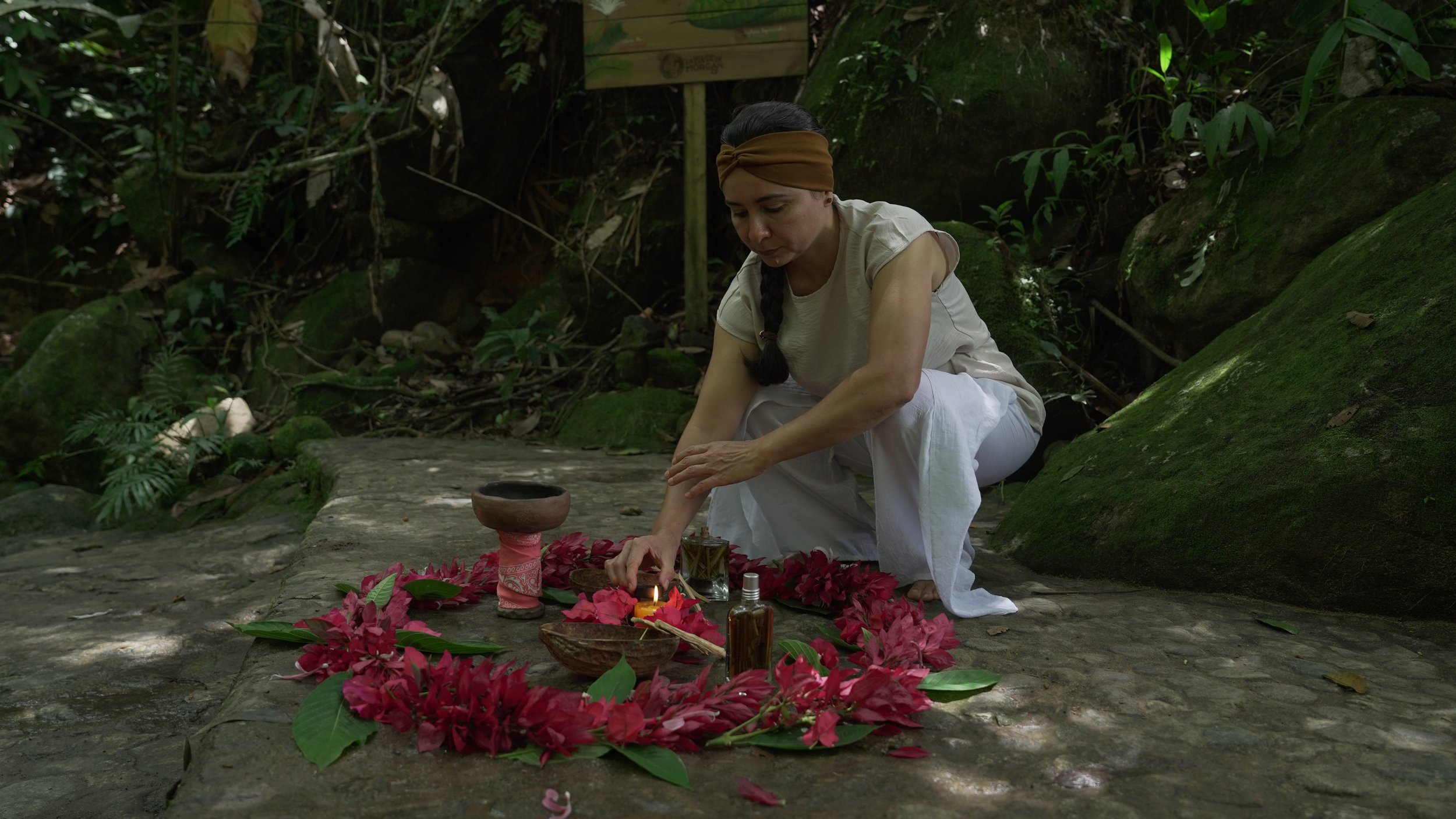 Mujer en vestimenta blanco, con cinta en la cabeza, realizando un ritual espiritual o ceremonial en un ambiente natural y rodeada de flores rosadas y objetos rituales.