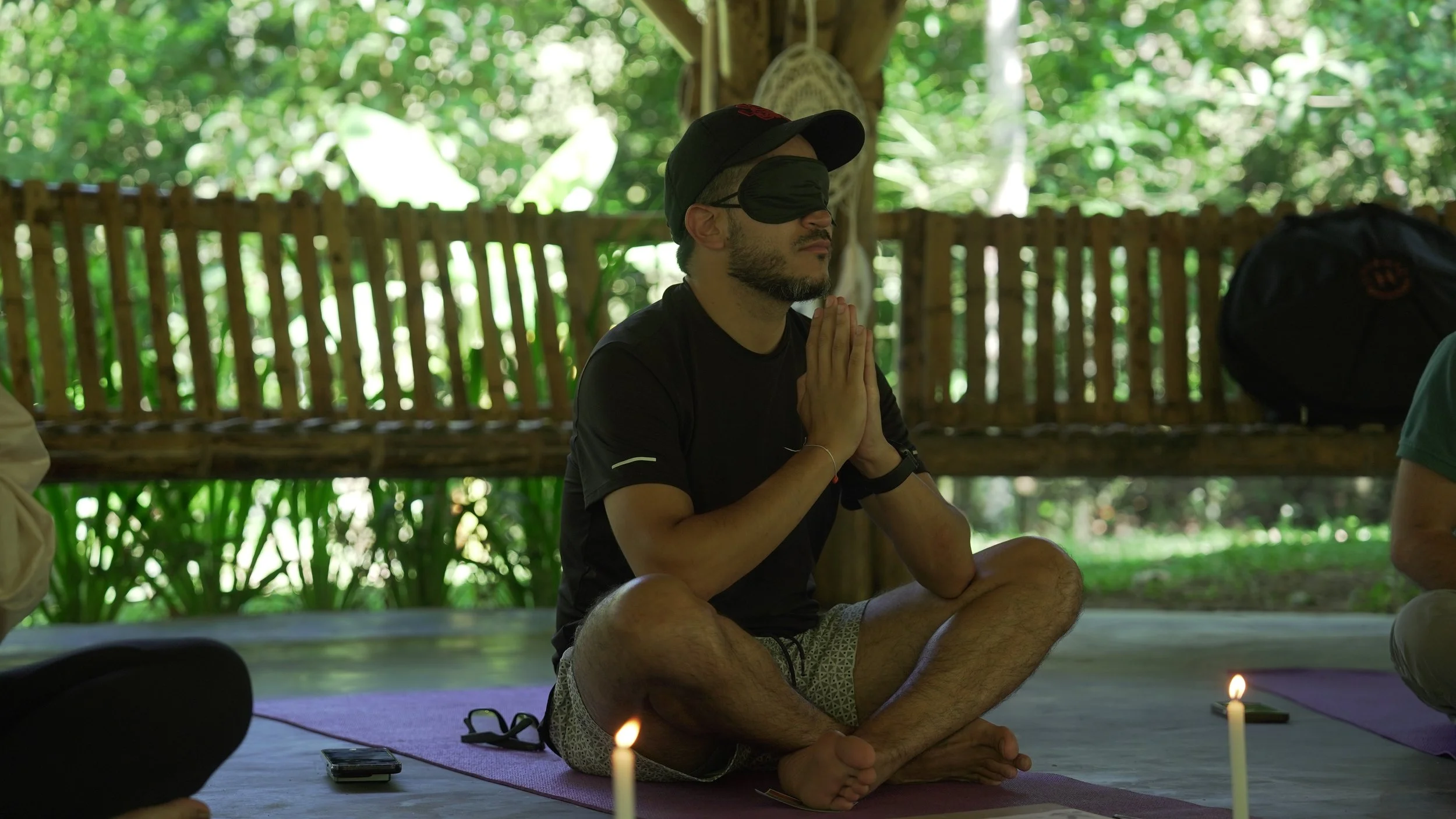 Hombre con gorra y ojos vendados, practicando meditación en plena naturaleza, en posición de loto, con velas encendidas alrededor.