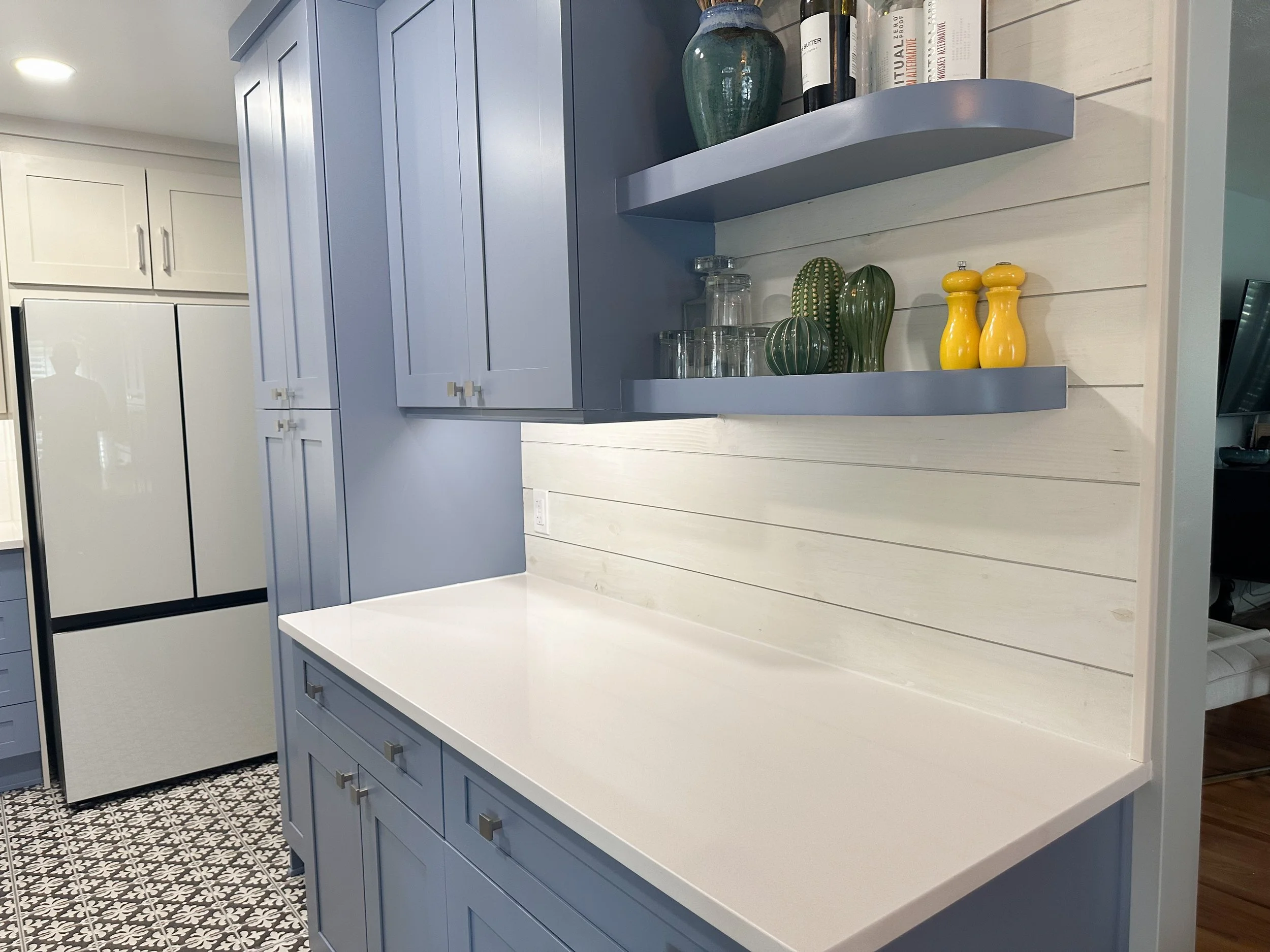 Modern kitchen with blue cabinets, a sleek white countertop, and floating shelves displaying decor items including green vases, glassware, books, and yellow salt and pepper shakers. The room features a patterned black and white tile floor and a large, light-colored refrigerator.