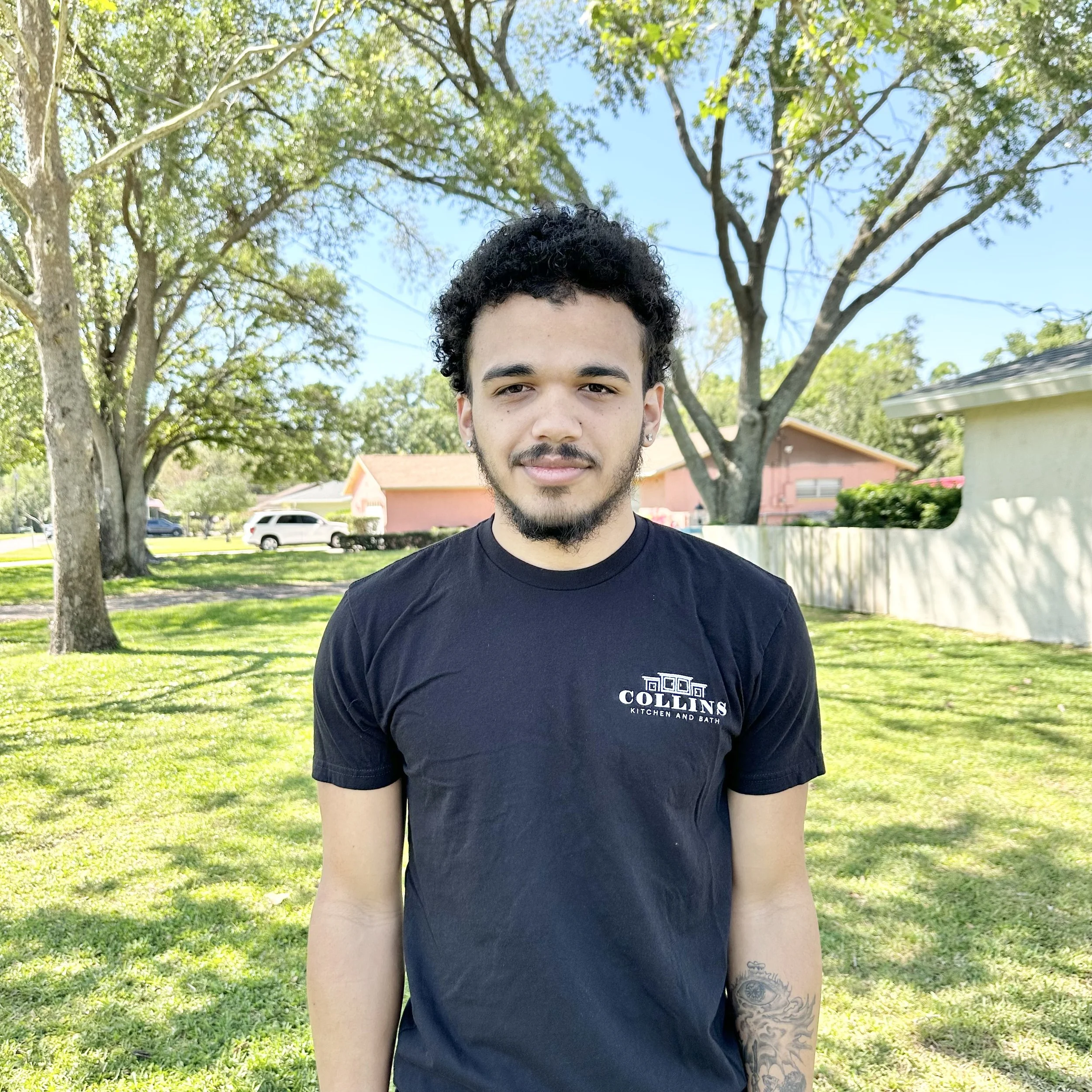 A person wearing a black T-shirt with "Collins Kitchen and Bath" logo, standing outdoors on a sunny day with trees and a house in the background.