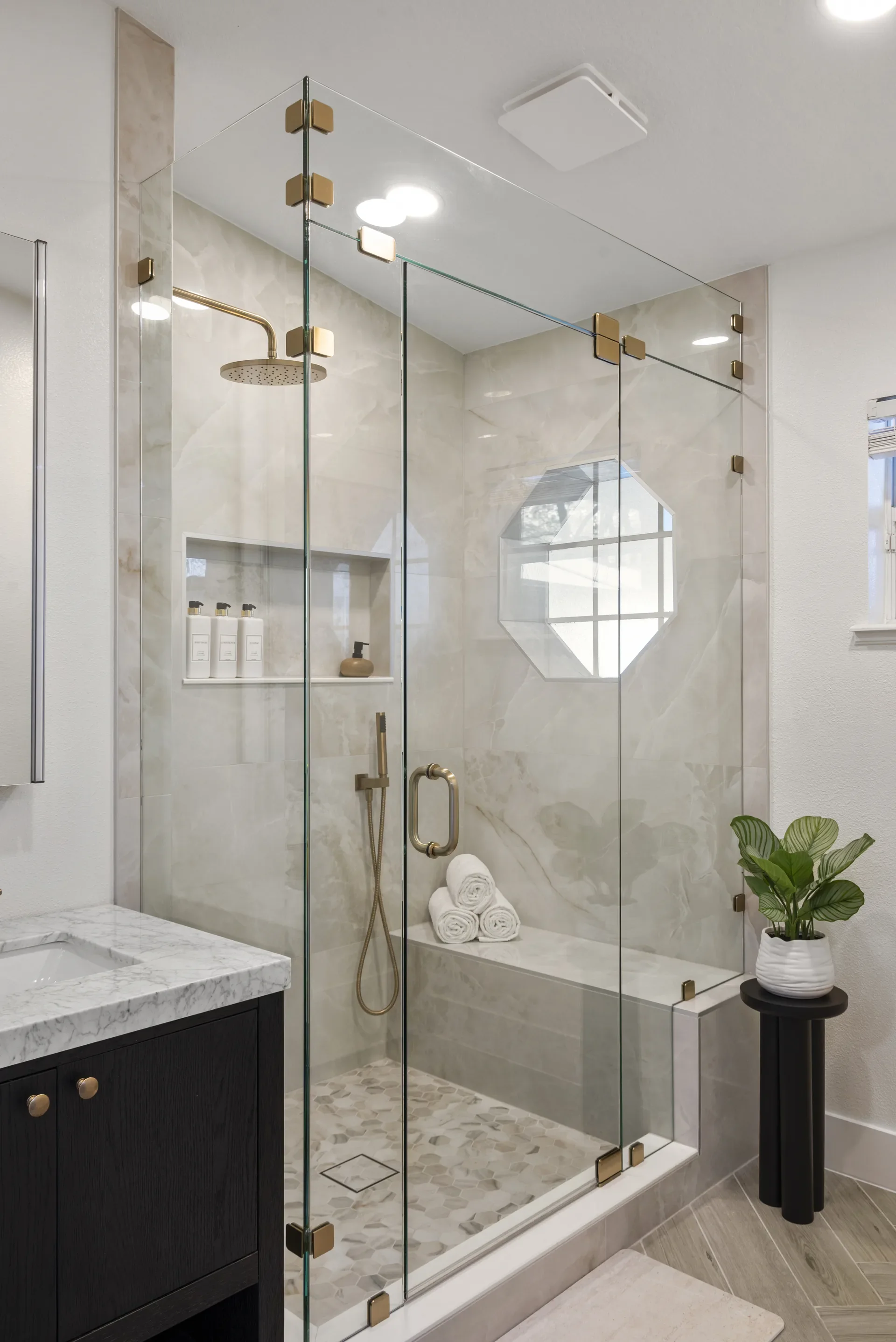 Modern bathroom with a glass shower enclosure, beige marble walls, gold fixtures, a marble vanity, towels, and a potted plant.