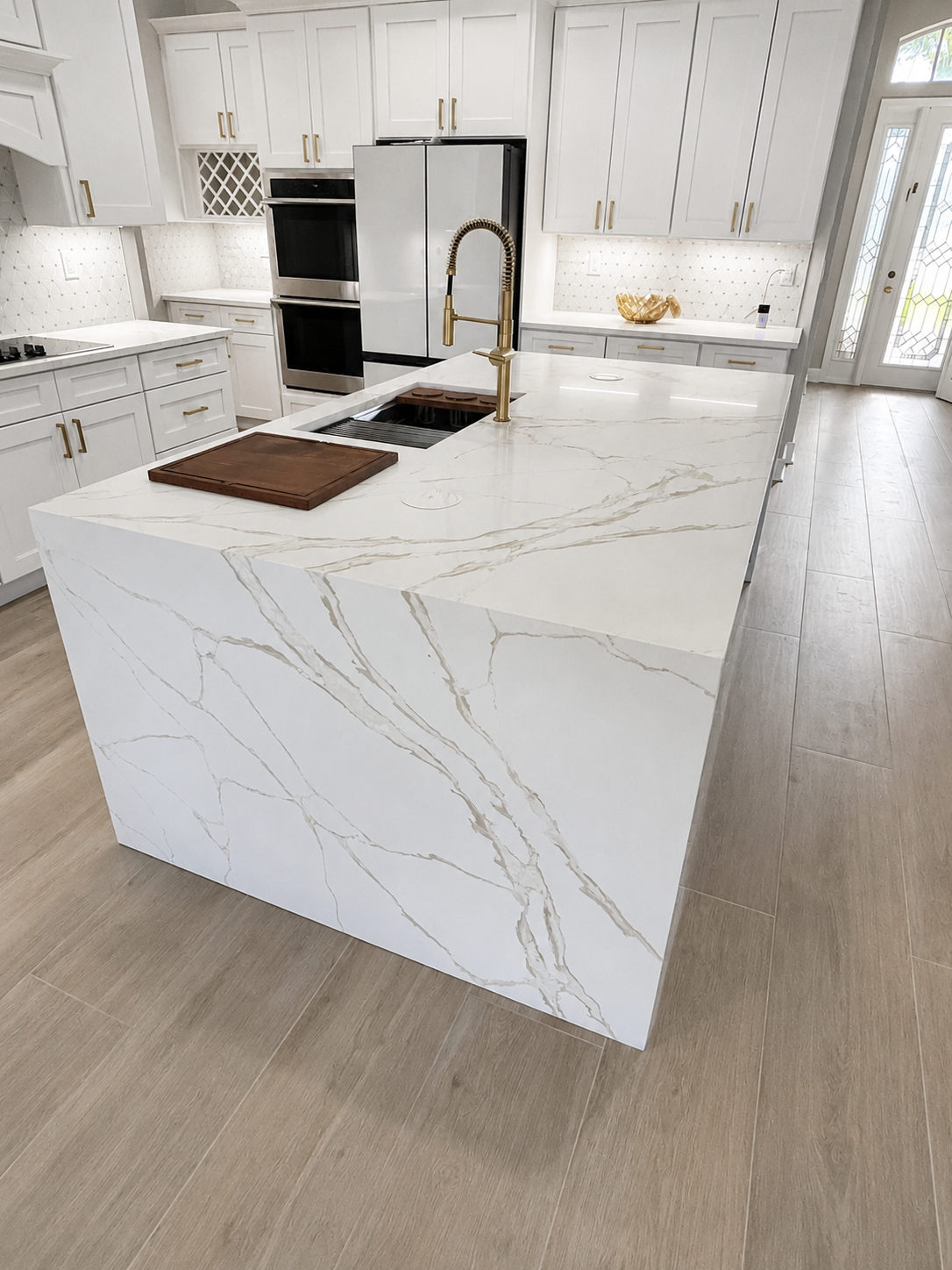 A modern kitchen with white marble island, gold fixtures, white cabinetry, and stainless steel appliances.