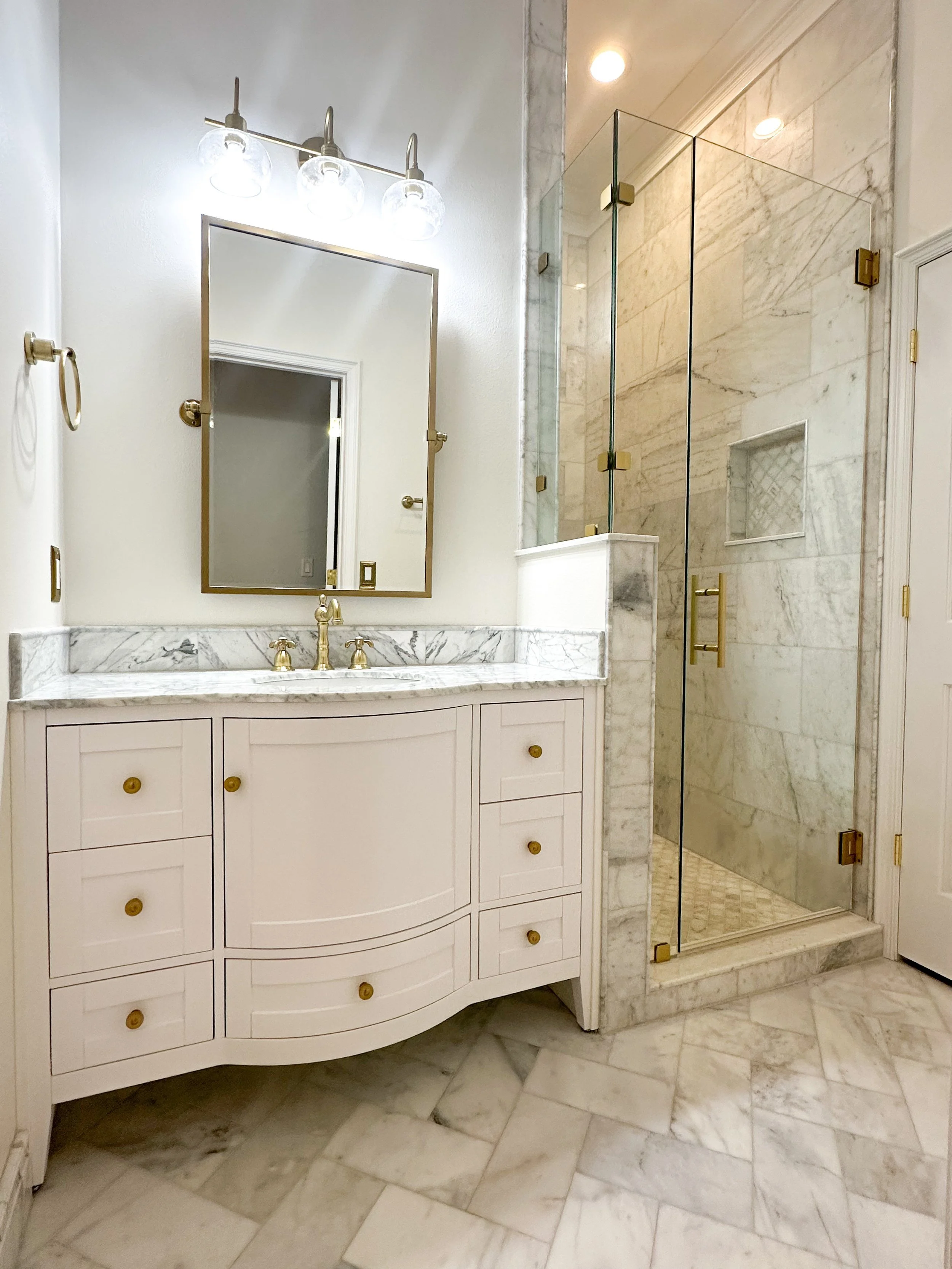 Bathroom remodel with curved white vanity, marble countertop, brass fixtures and a glass walk-in shower