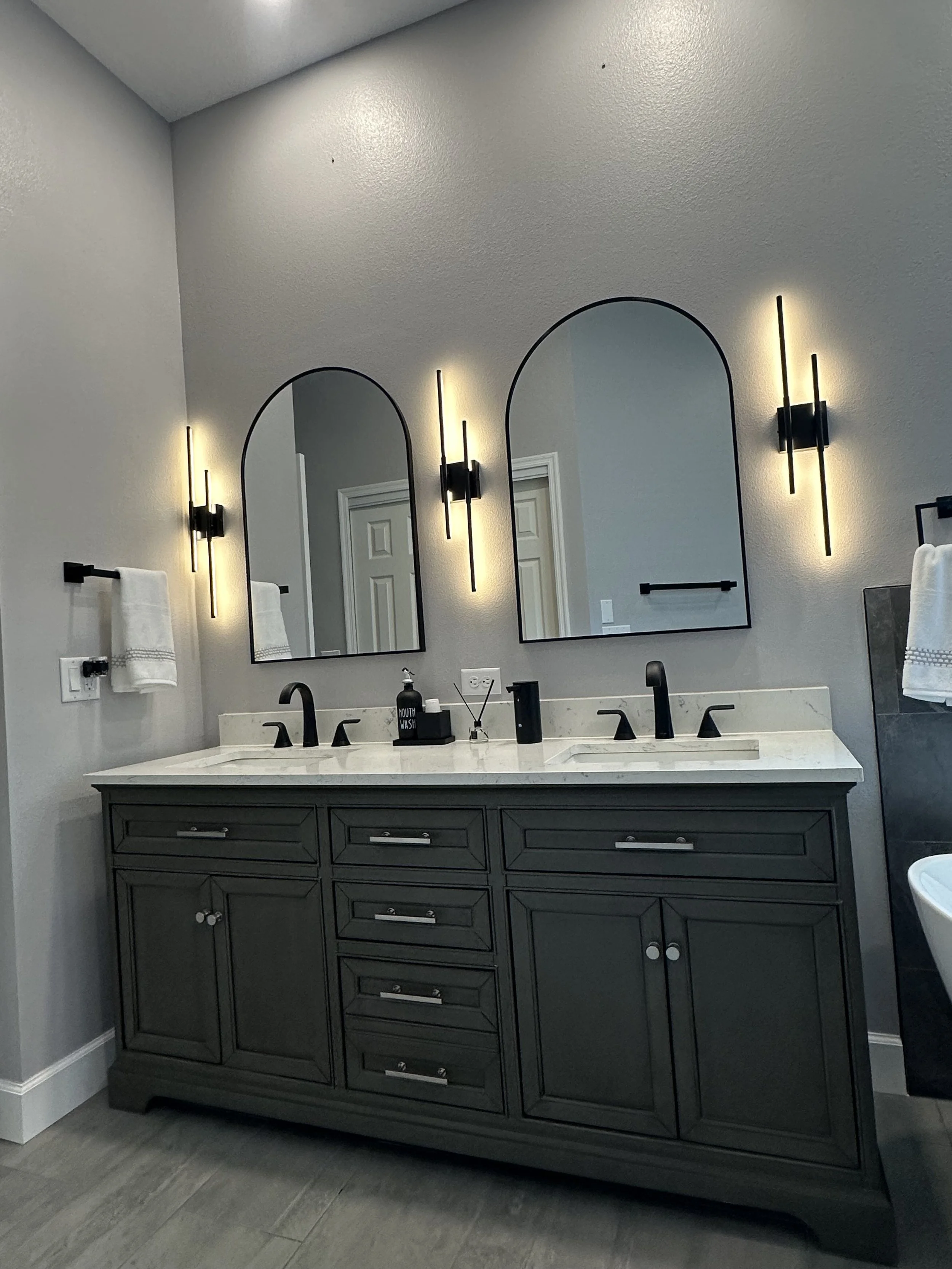 Charcoal double vanity with white quartz, arched black mirrors, and lit linear sconces in a Heathrow, Lake Mary primary bath.