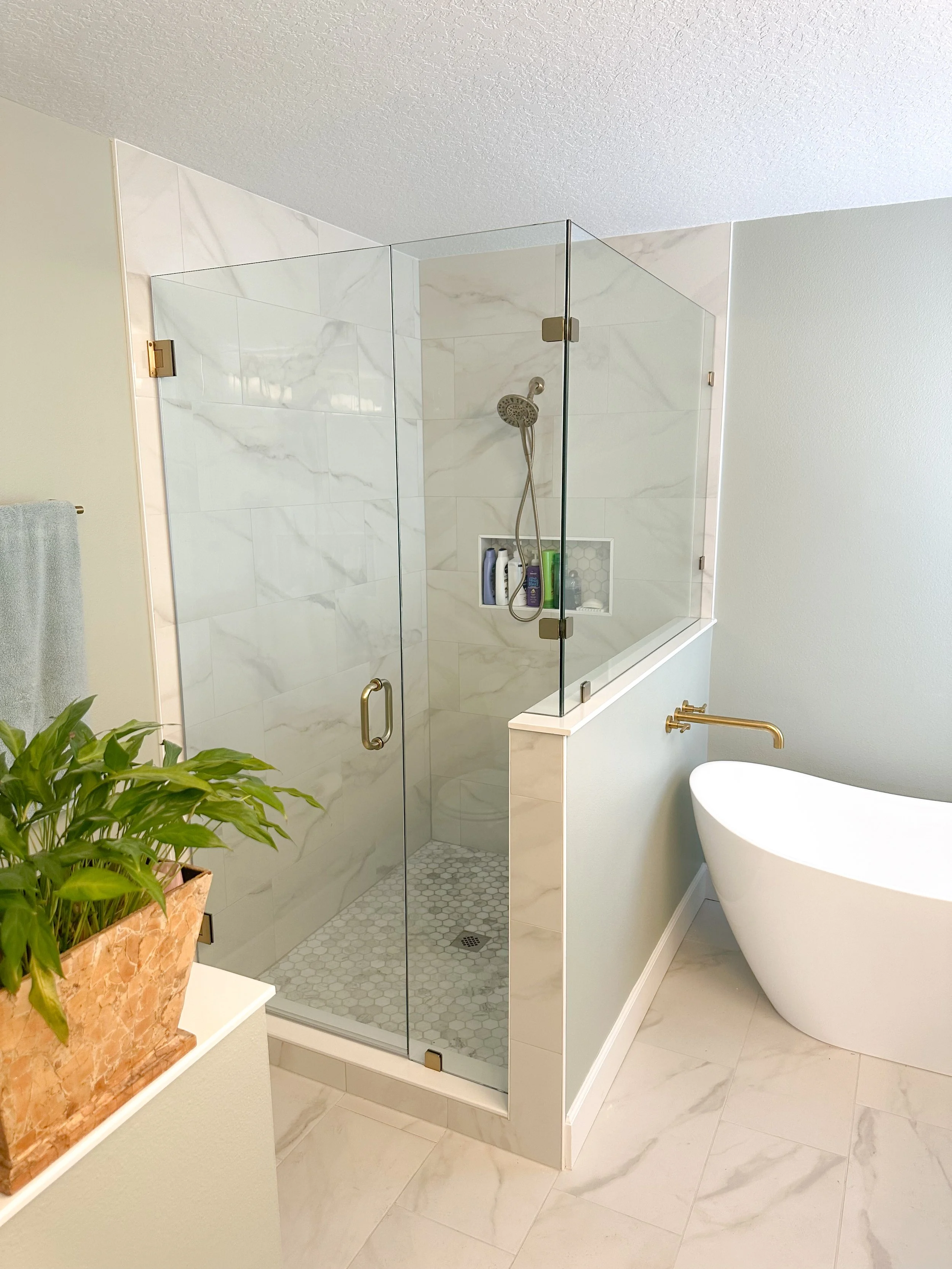 Modern bathroom with a glass-enclosed shower, a freestanding tub, marble tile, brass fixtures, and a potted plant.