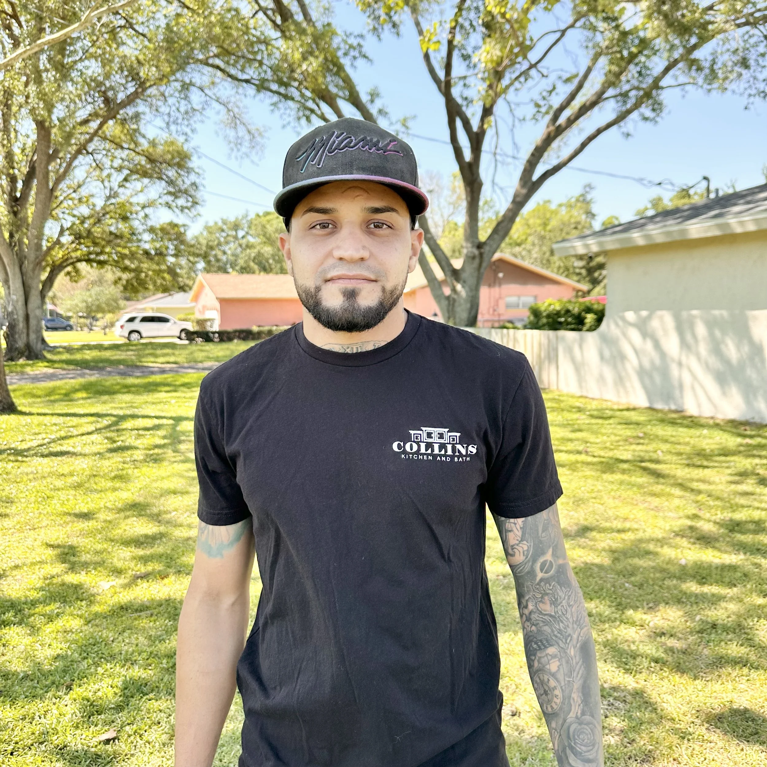 Man wearing a black "Collins Kitchen and Bath" t-shirt and a Miami cap standing on a grassy lawn with trees and houses in the background.