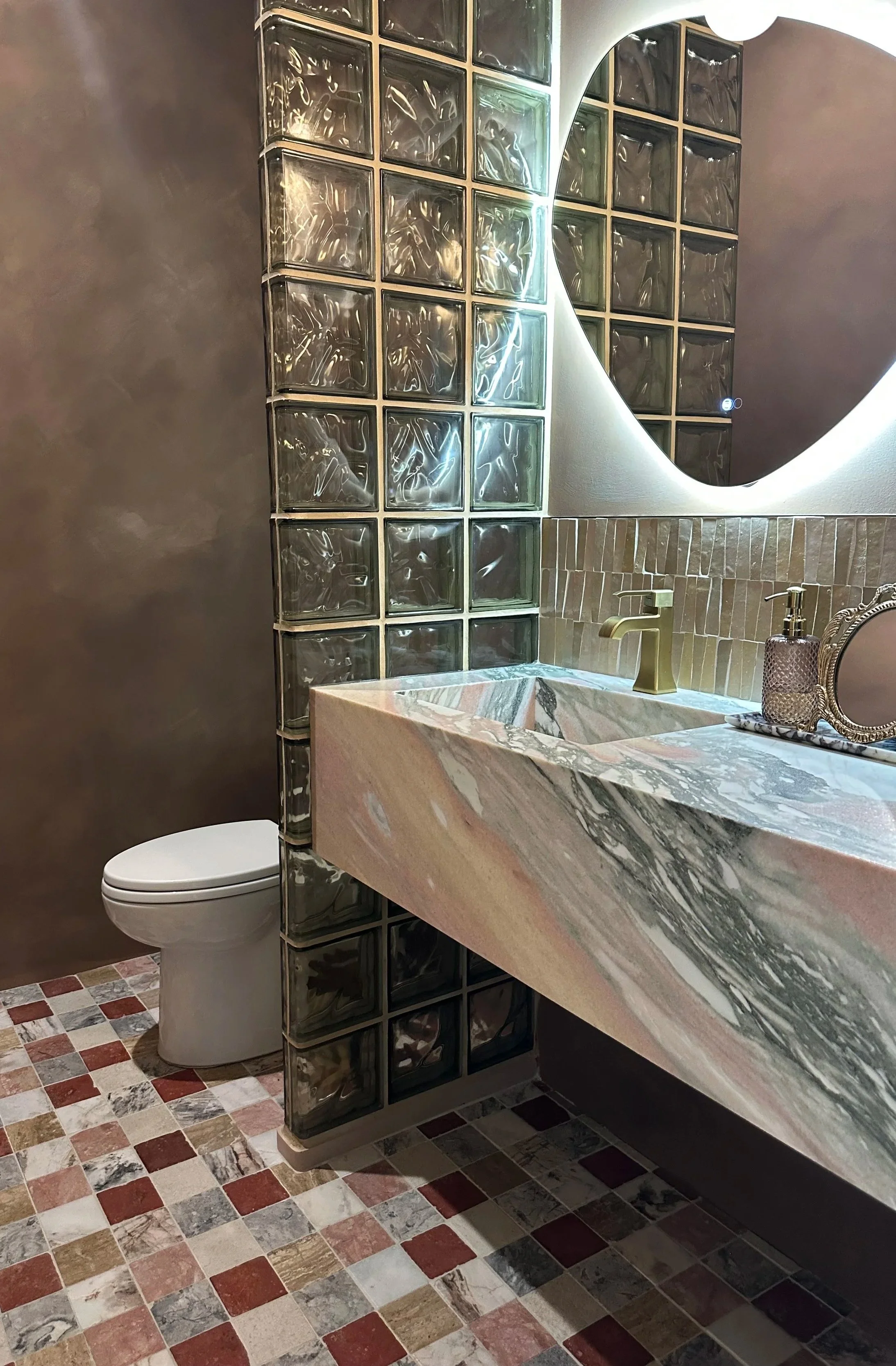 A bathroom featuring a glass block wall, a marble sink countertop with a gold faucet, a decorative soap dispenser, a mirror with a rounded corner, a toilet, and multicolored tile flooring.