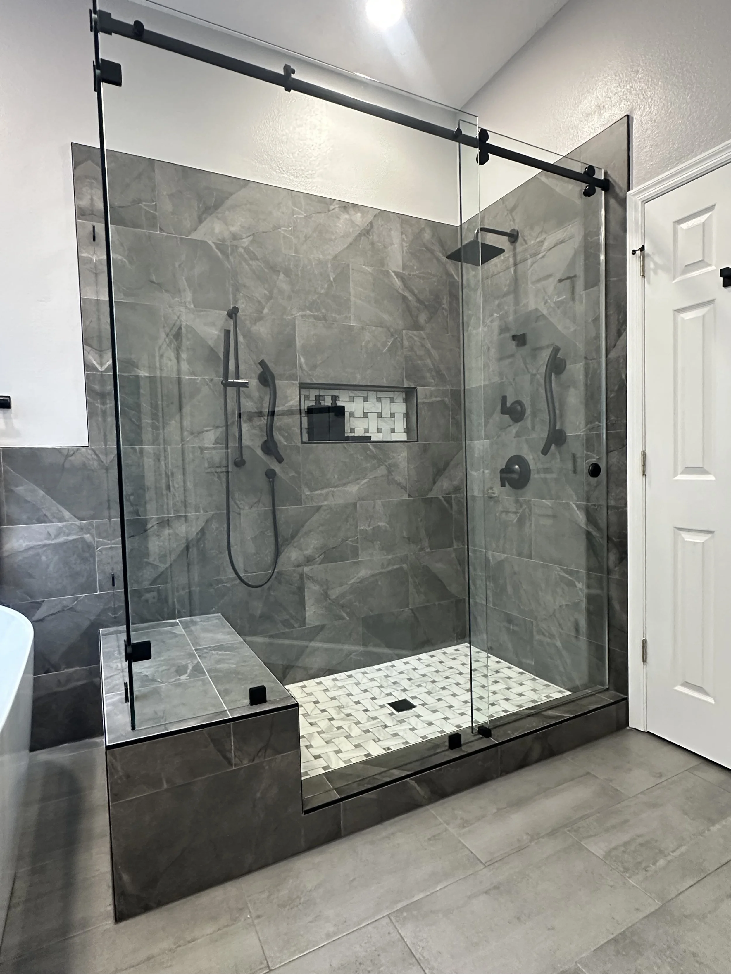 Walk-in shower with frameless glass, gray marble-look tile, a tiled bench, and a basketweave marble floor in a Lake Mary primary bath.