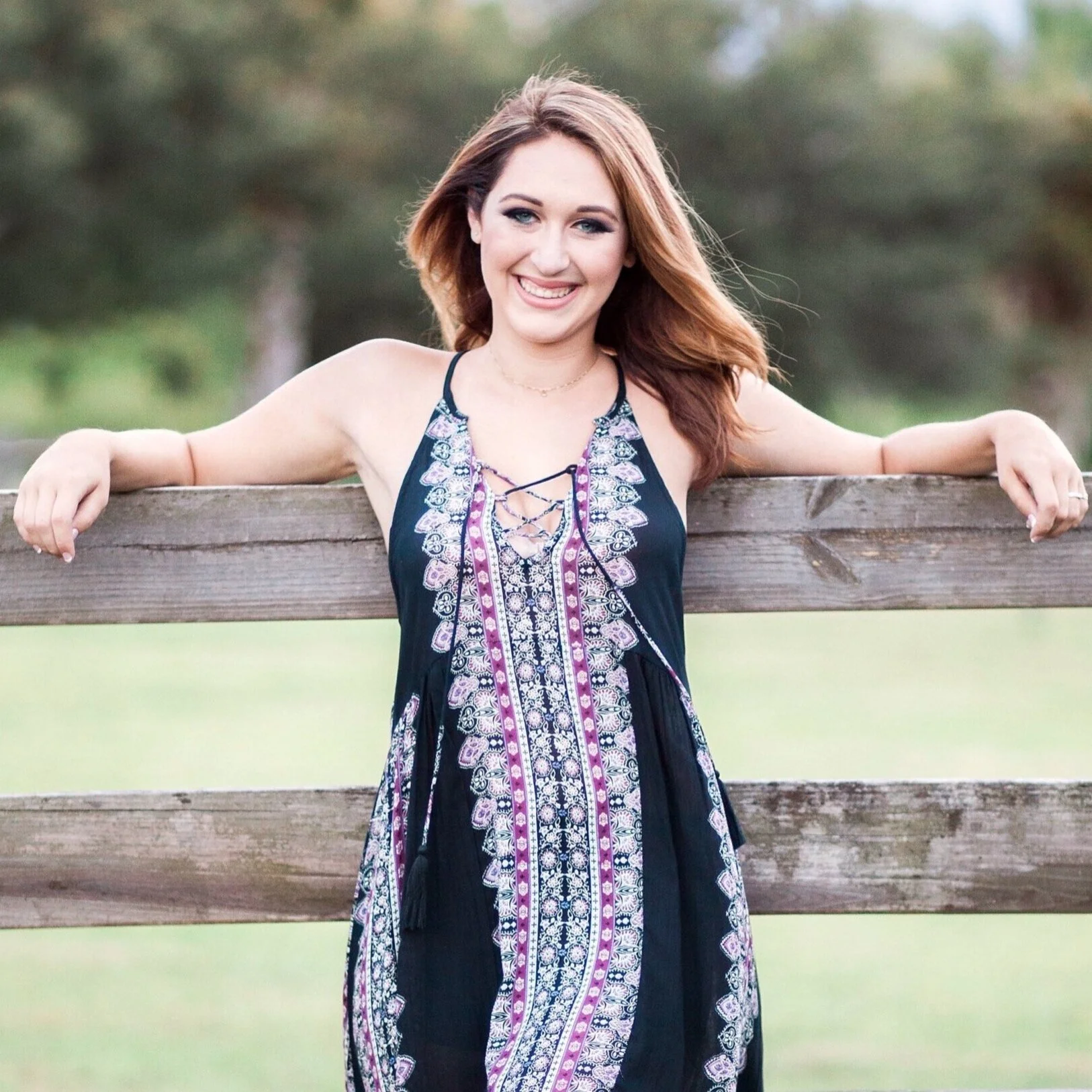 Woman in patterned dress smiling, leaning on a wooden fence with a blurred green background.