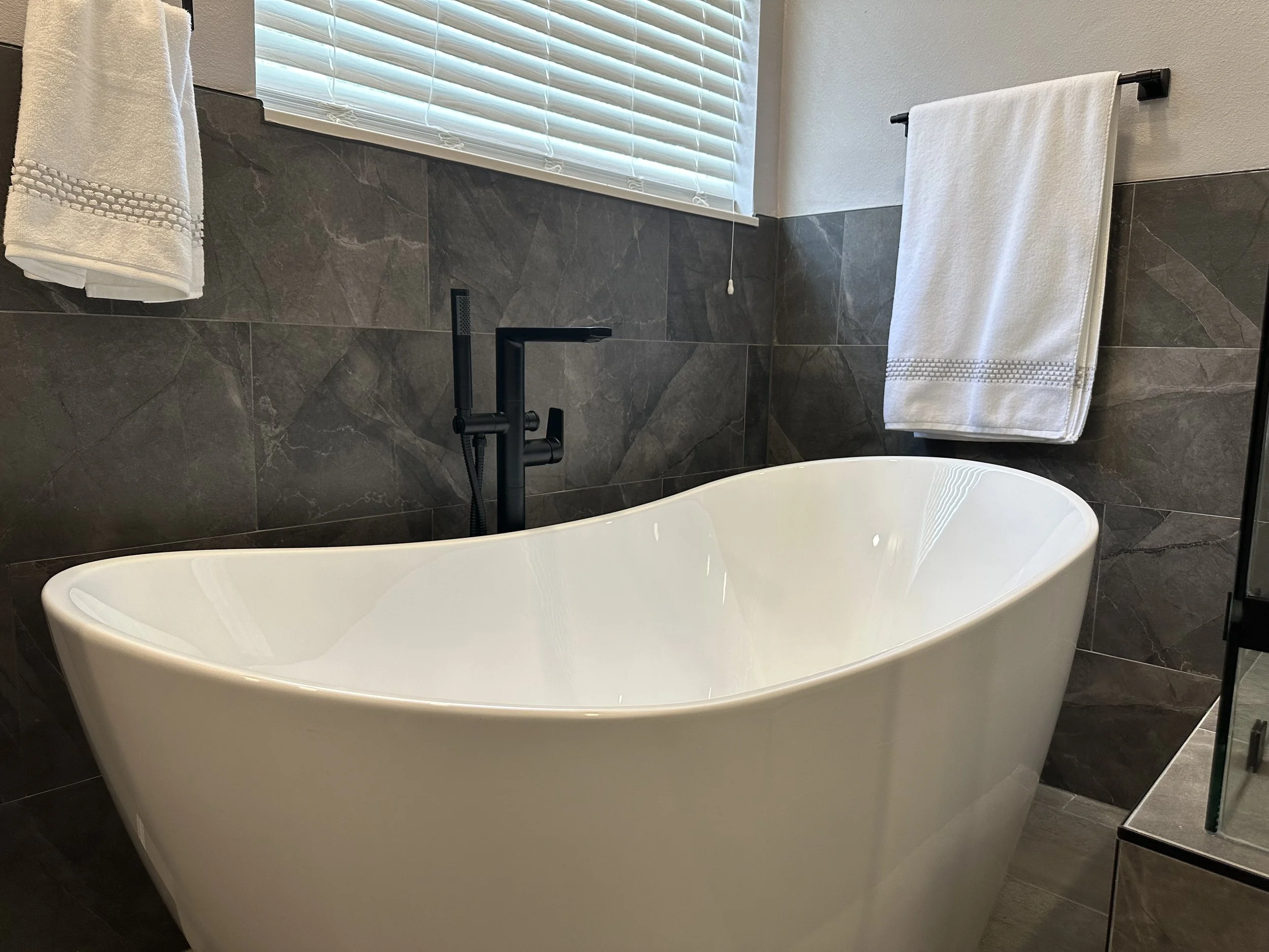Freestanding soaking tub with a matte black floor-mounted tub filler against gray marble-look tile in a Heathrow primary bathroom.