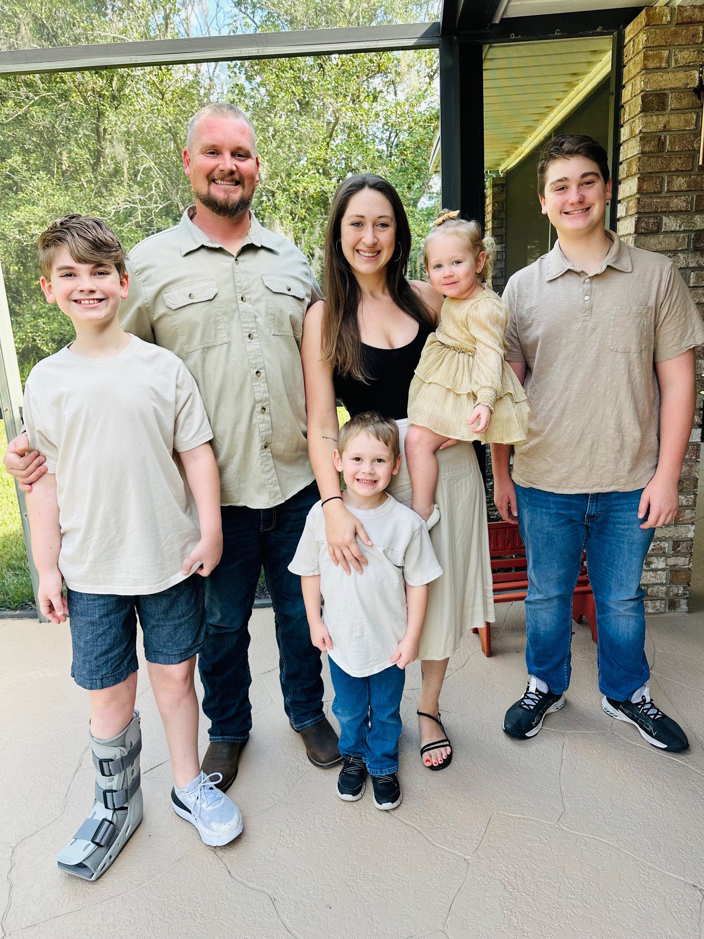 A family of six posing for a photo on a patio. The group includes an adult man, woman, three young boys, and a toddler girl. One boy is wearing a medical boot. They're all smiling, wearing casual clothing, and standing in front of a brick wall with greenery in the background.
