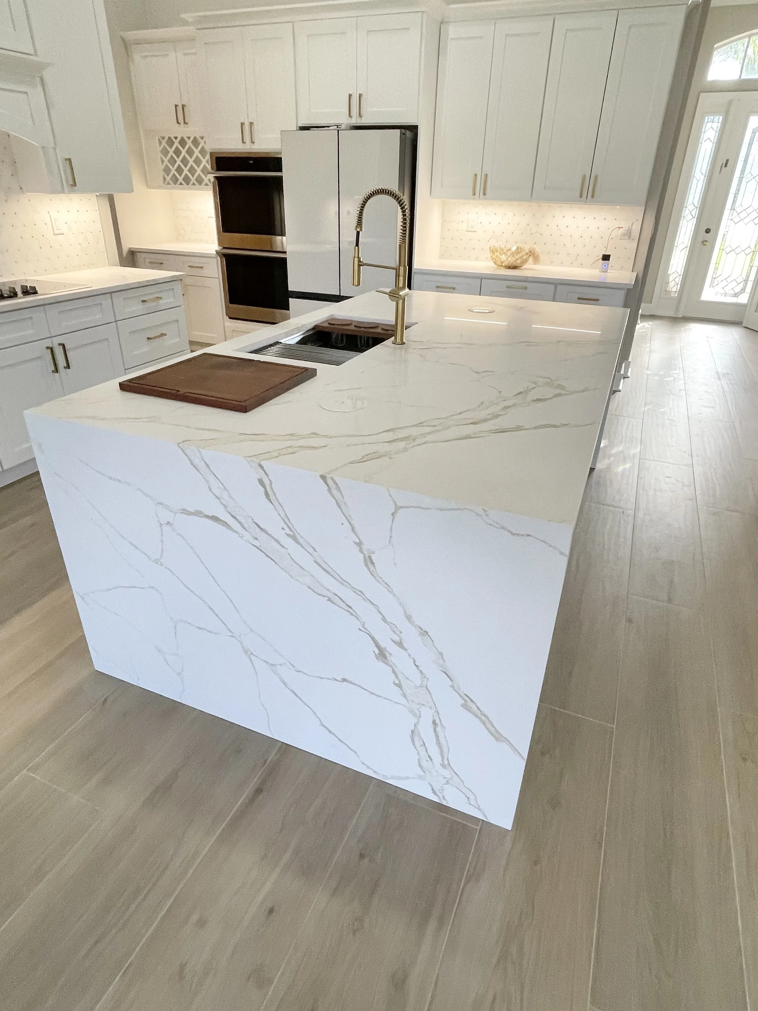 Modern kitchen featuring a large marble island with a gold faucet, white cabinets, and hardwood flooring. Includes built-in double ovens and a stainless steel refrigerator.