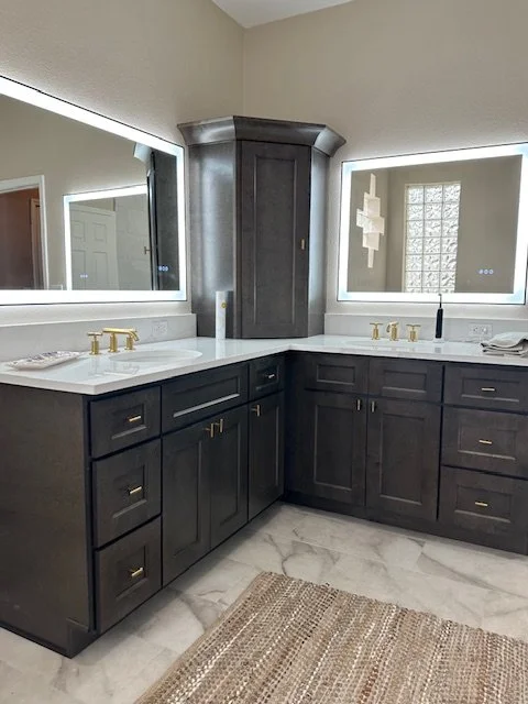 Modern bathroom with dark wood cabinets, white countertops, and integrated LED-lit mirrors. Features gold faucet fixtures and a woven rug on the marble floor.