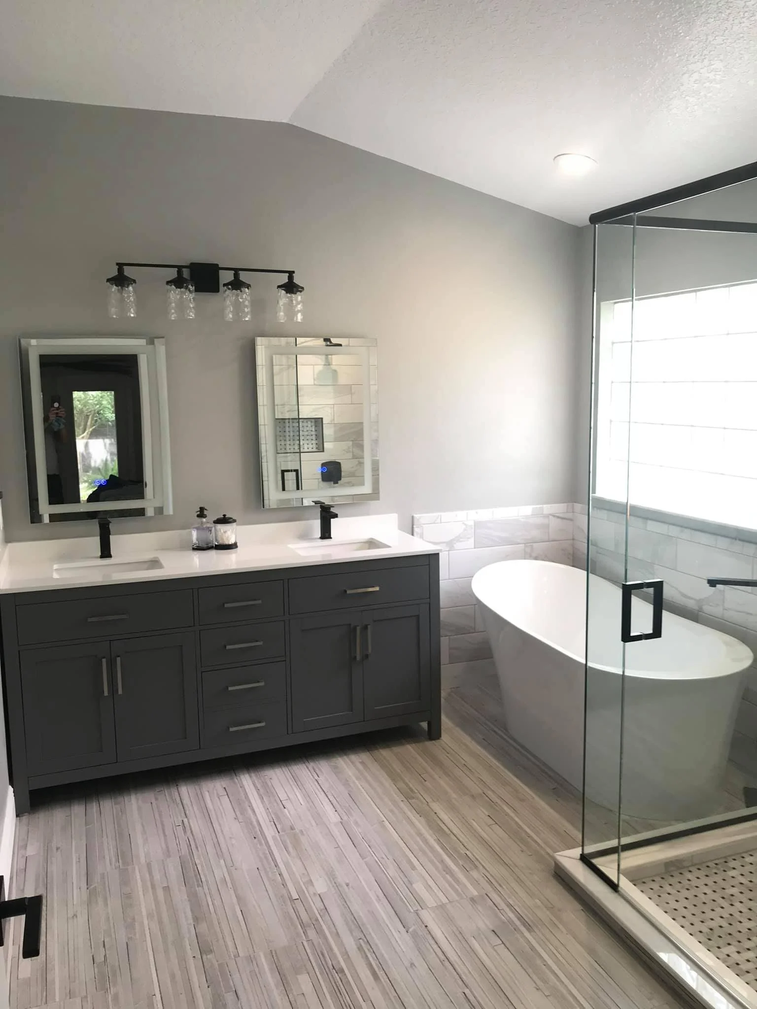 Modern bathroom with gray vanity, double sinks, round freestanding bathtub, glass shower enclosure, and tiled flooring.