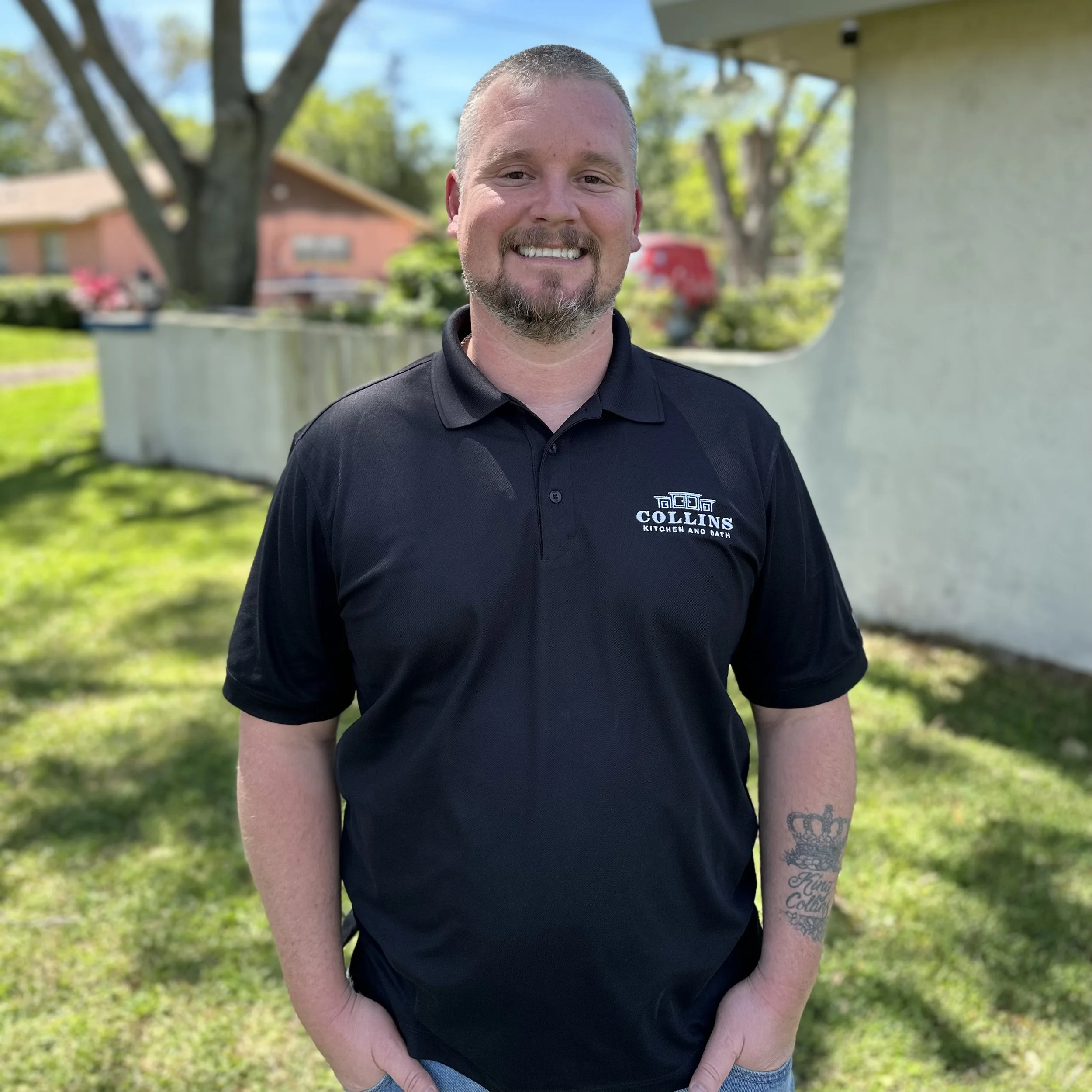 A man in a black polo shirt stands outdoors with a house in the background.