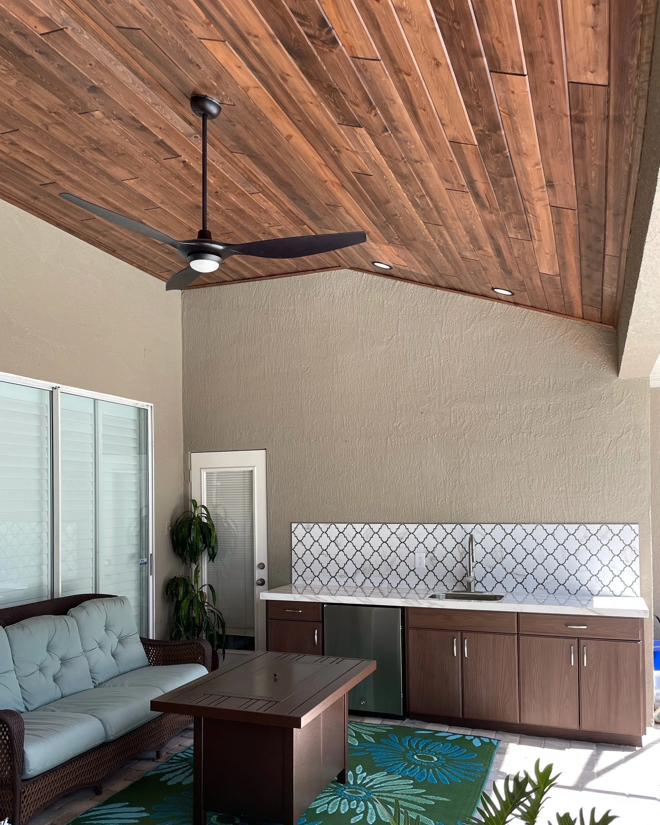 Outdoor patio with a wooden ceiling, a ceiling fan, a wicker sofa with cushions, a rectangular coffee table, a kitchenette with a sink, cabinets, and a patterned tile backsplash, sliding glass doors, and decorative plants.