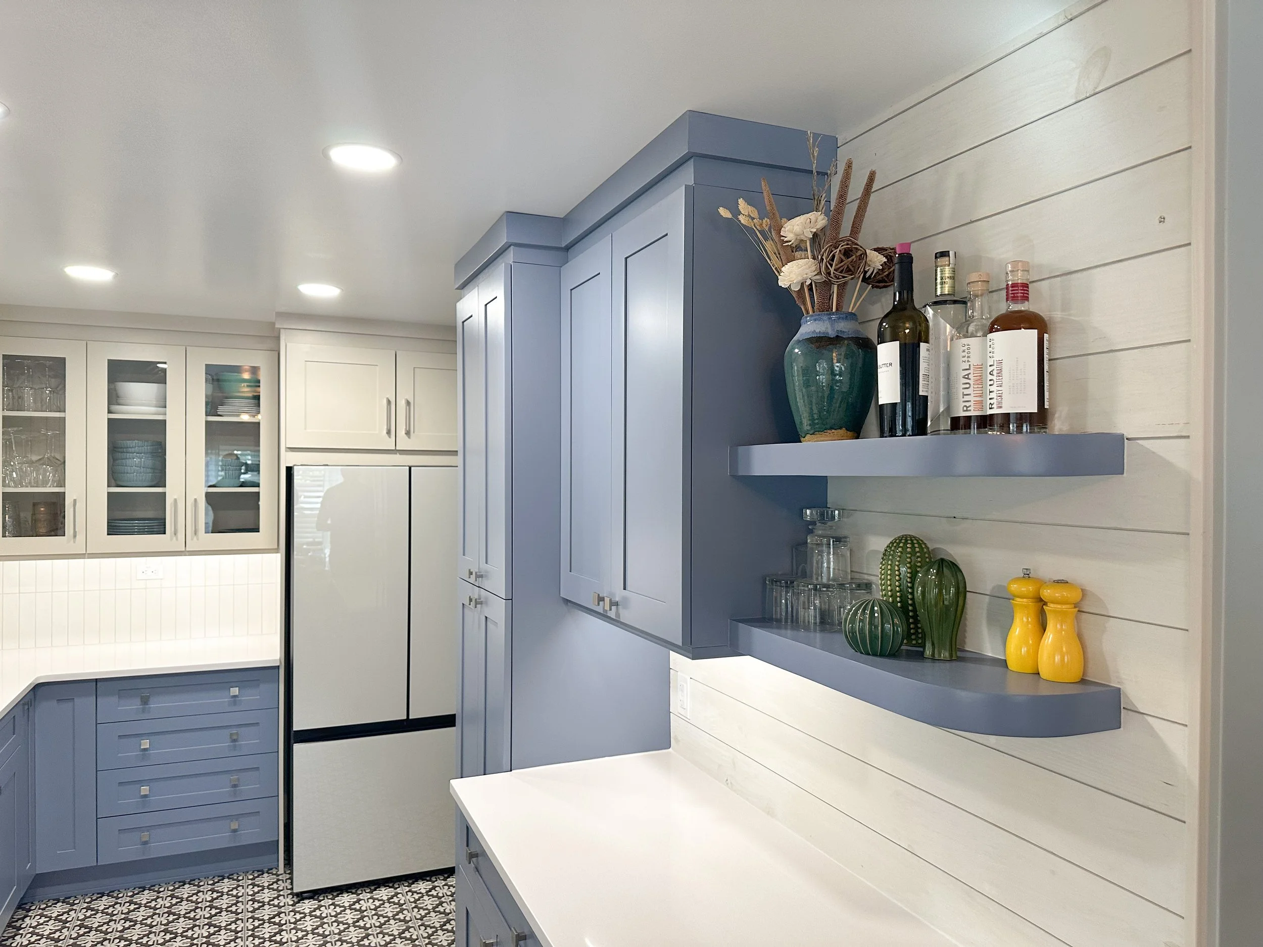 Modern kitchen with blue cabinets, white countertops, and patterned tile flooring. Open shelves display a green vase, bottles, glassware, and decorative items. A refrigerator is integrated into the cabinetry.
