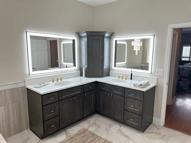 Bathroom with double vanity, large LED-lit mirrors, dark wooden cabinets, white countertop, brass faucets, and marble floor tiles.