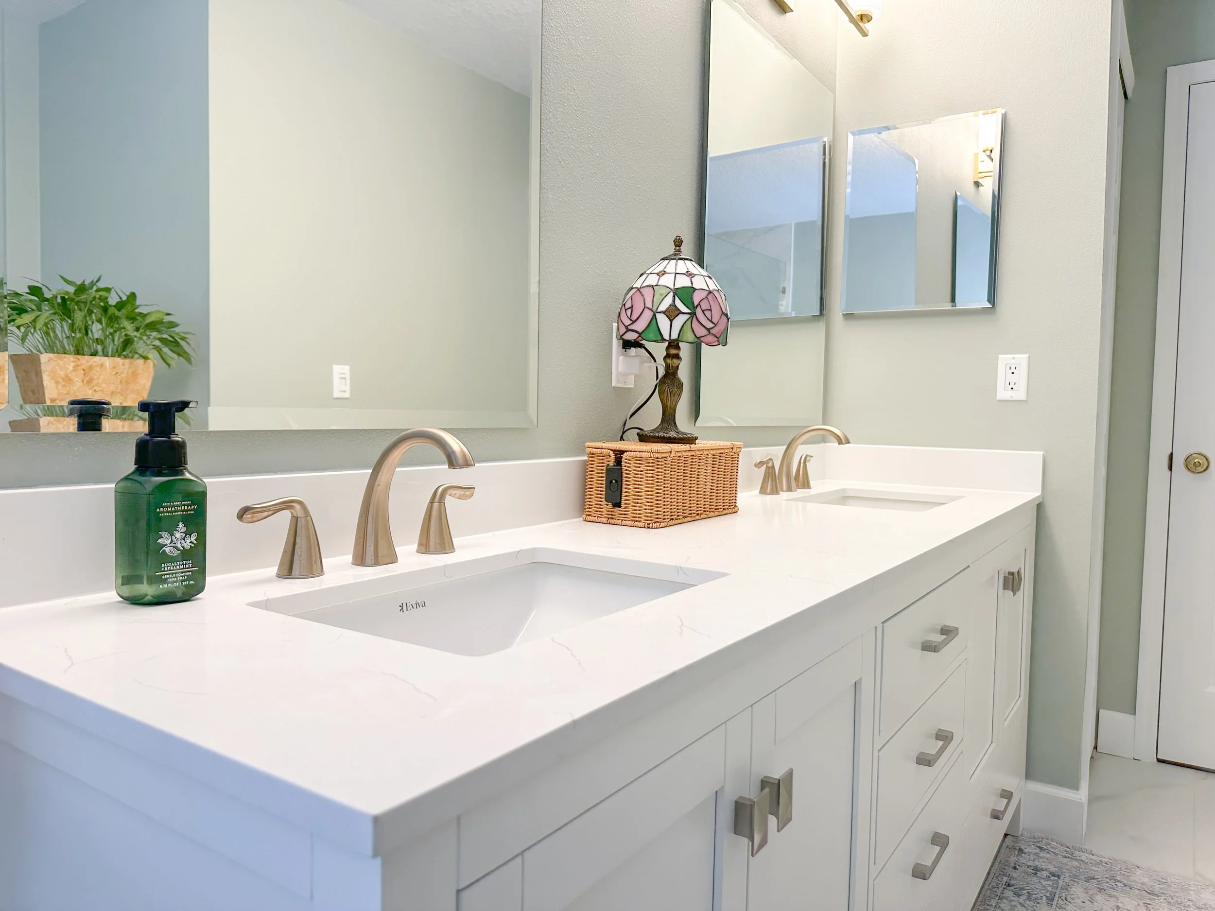 Modern bathroom countertop with dual sinks, gold faucets, a decorative lamp, wicker basket, green plant, and soap dispenser in a bright setting.