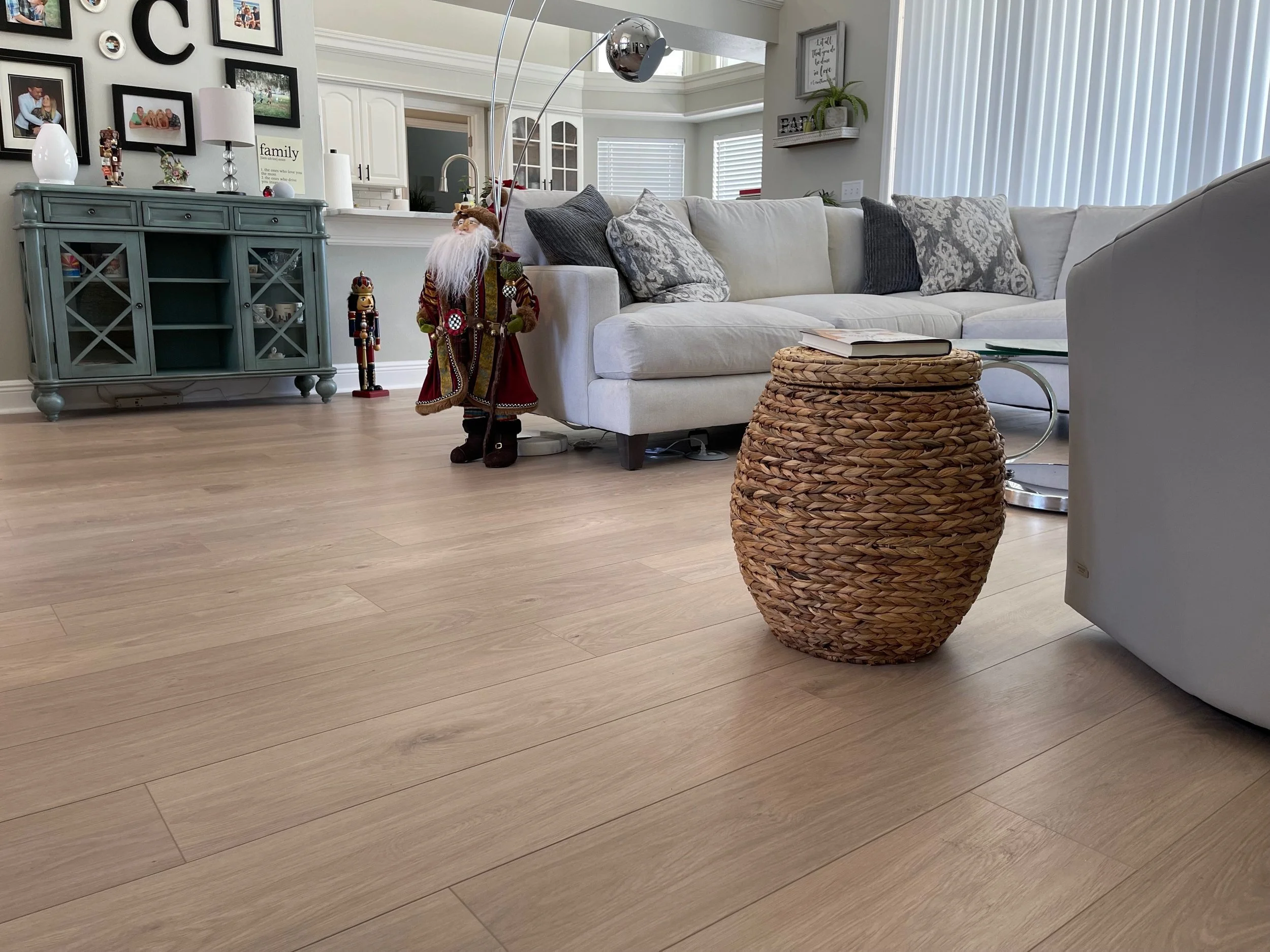 Living room with light wood flooring, a white couch with decorative pillows, a woven side table, a blue-green cabinet against the wall with family photos and "C" decor. Santa figurine in festive attire and a nutcracker decoration beside the cabinet. Floor lamps and vertical blinds in the background.