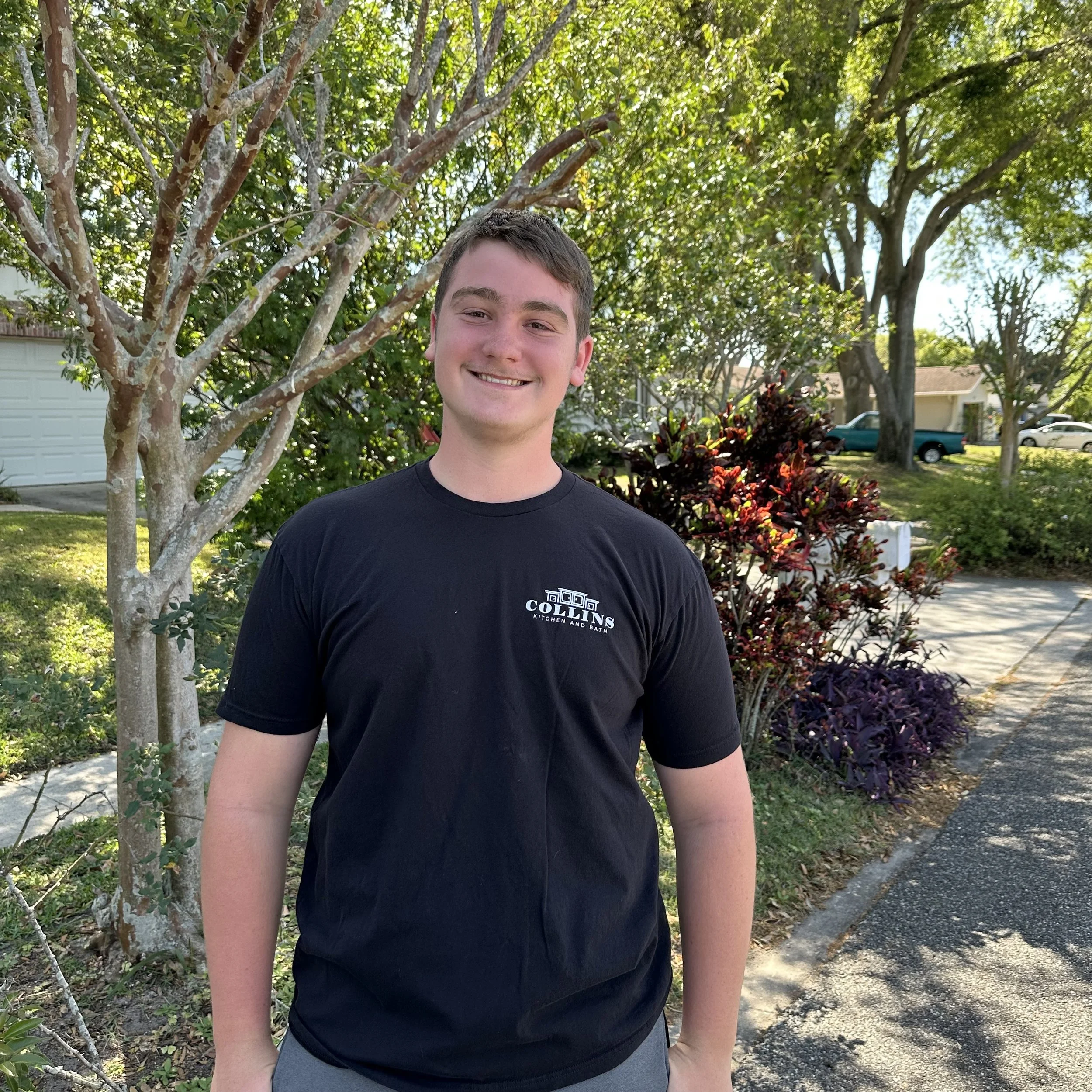 Person smiling in a black t-shirt standing outdoors near trees and shrubs on a sunny day.