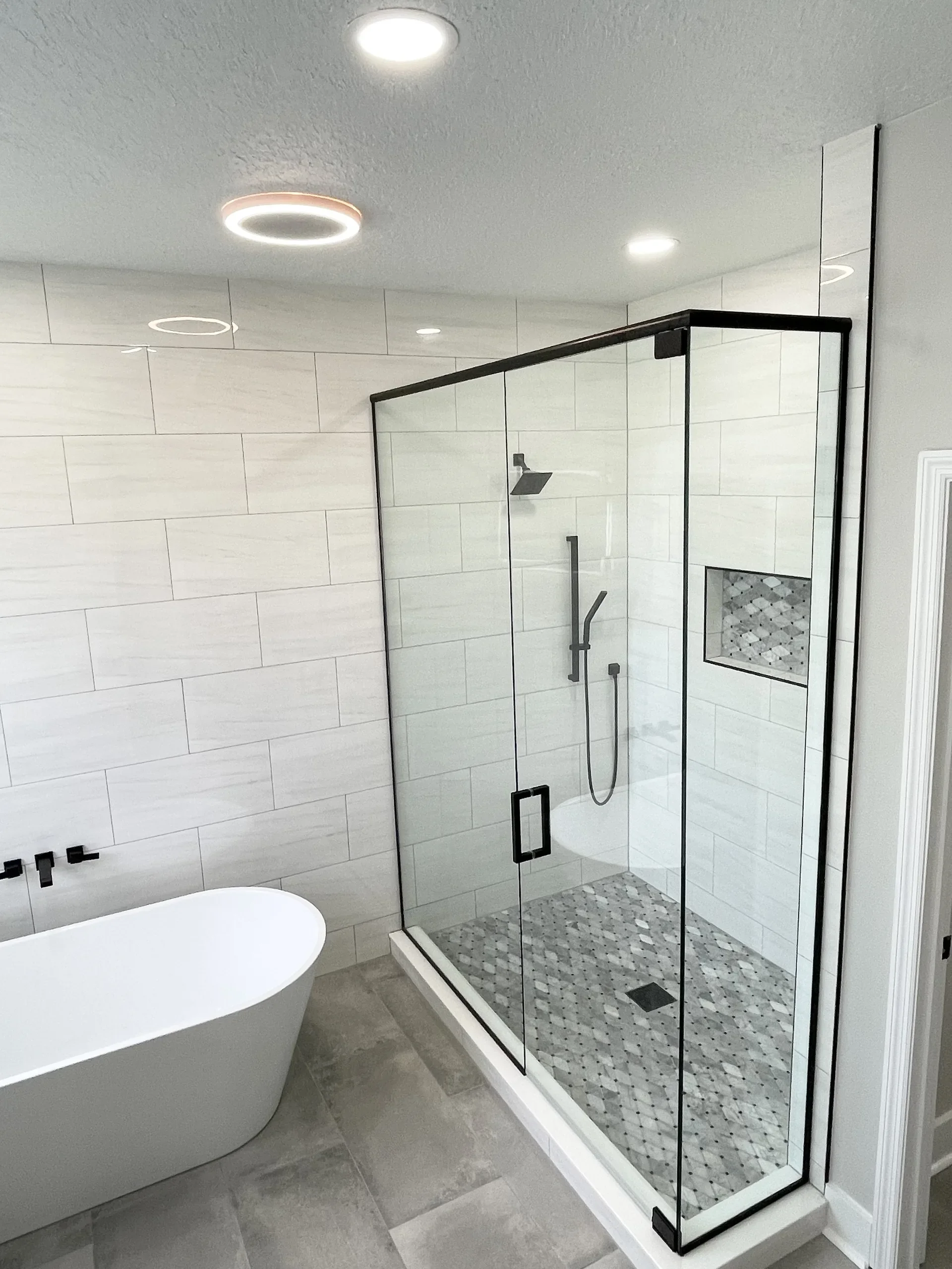 Modern bathroom with a freestanding bathtub, a glass shower enclosure with black framing, and light gray tiled walls and floor.