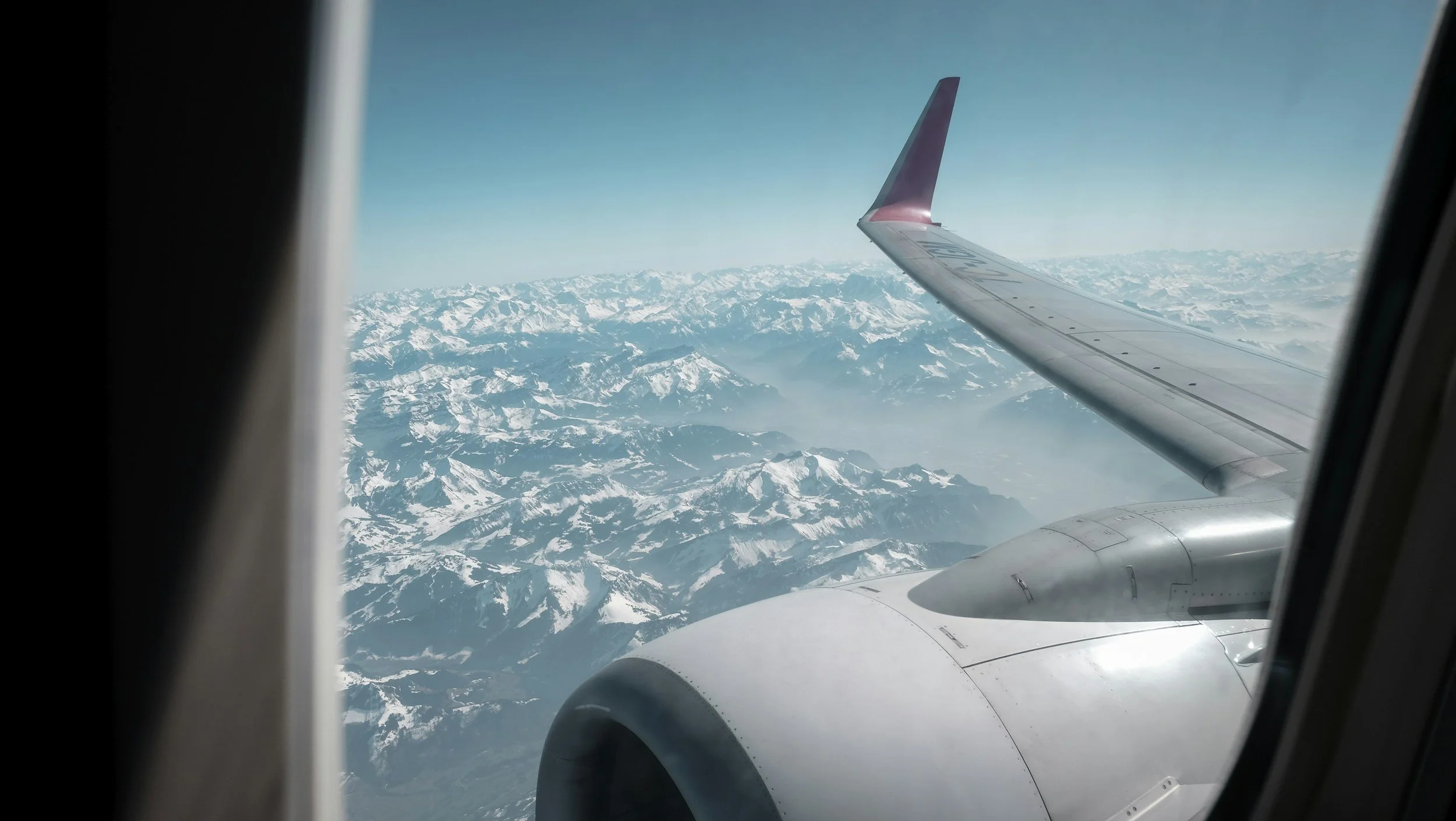 View of snowy mountains from an airplane window, showing the plane's wing and engine.