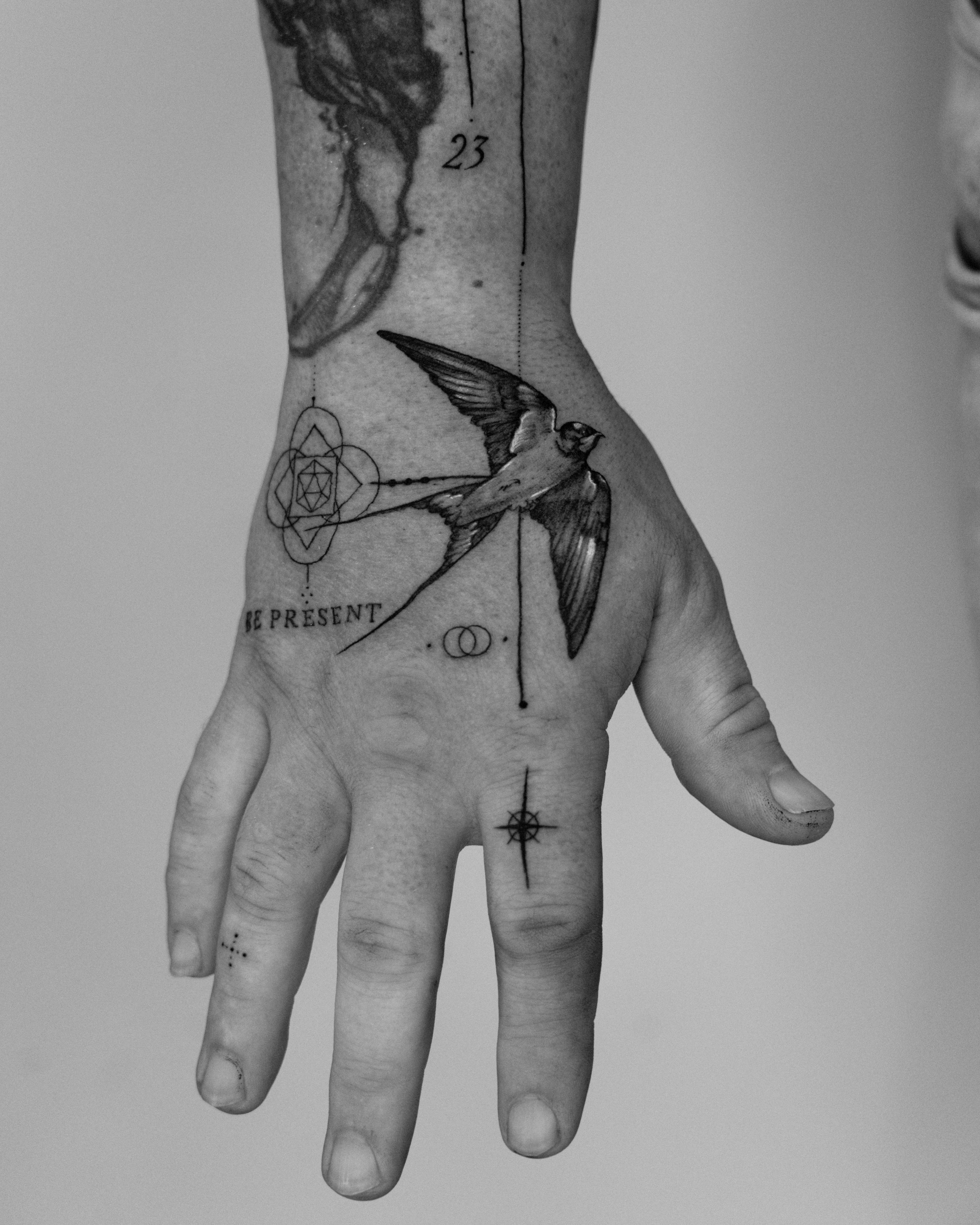 Micro realism tattoo in Brighton of a tattooed hand and wrist with various black ink tattoos, including a bird in flight, geometric shapes, and words "BE PRESENT." The image is in black and white.
