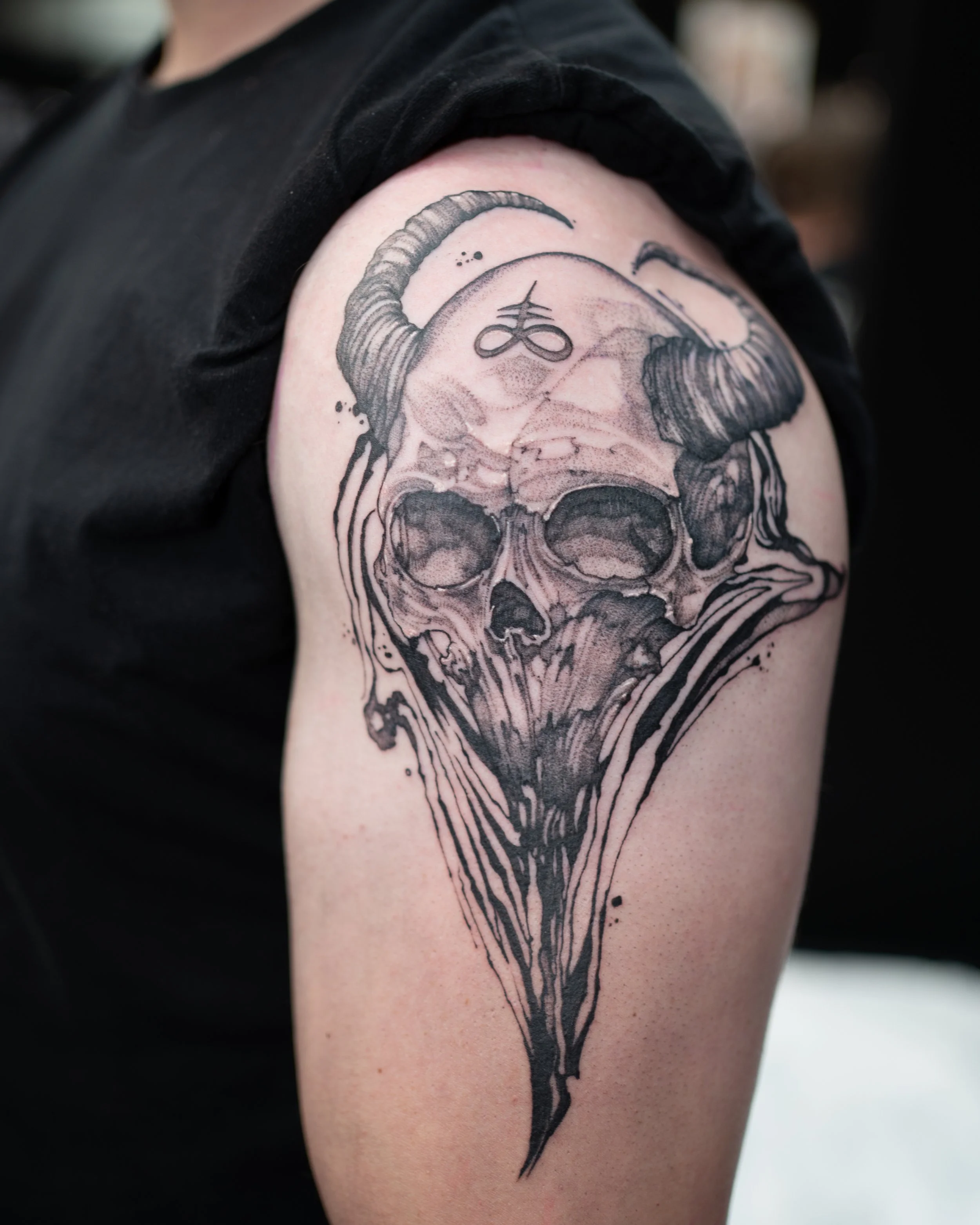 Black and gray tattoo of a skull with ram horns, a Satanic symbol on the forehead, and dark dripping lines extending downward.