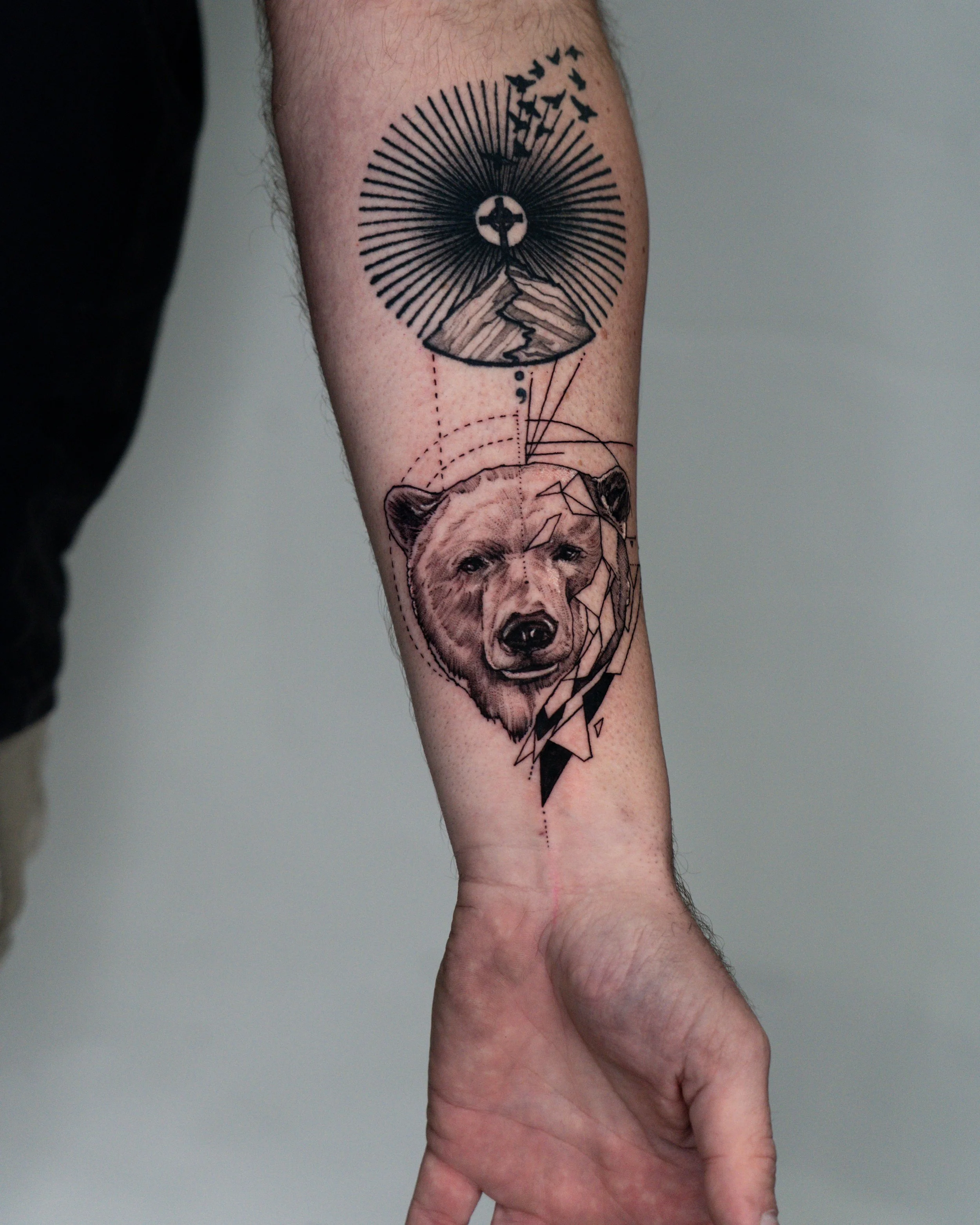 Brighton tattoo artist creates a piece on forearm featuring a bear's head, a mountain landscape, and abstract geometric elements with lines and dots.