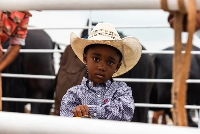 A young boy wearing a cowboy hat and a button-up shirt stands behind a fence at a cattle farm.