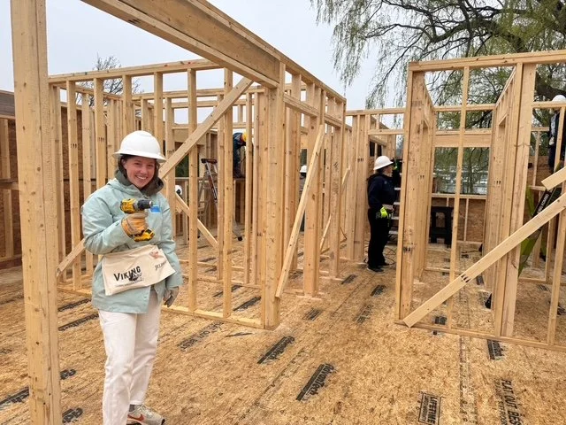 Women wearing construction helmets and gloves working on framing a house under construction, with wooden beams and plywood flooring.