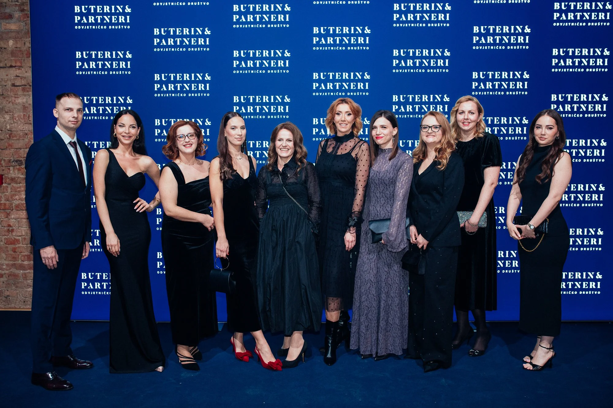 Group of eleven people, mostly women, dressed in formal attire, standing in front of a blue backdrop with white text that reads 'BUTERIN & PARTNERI' and 'ODVJETNICKO DRUŠTVO', at a formal event or gathering.