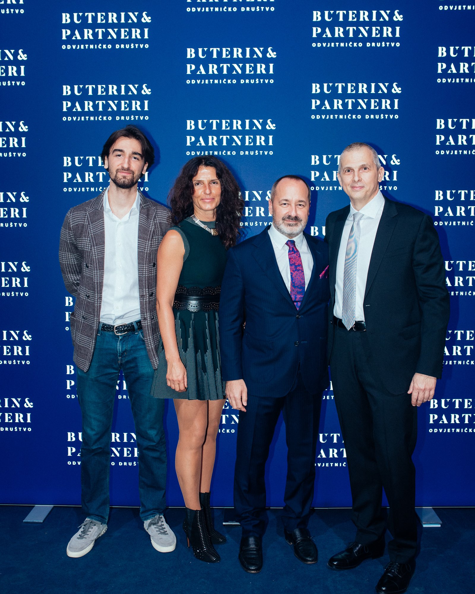 Four people standing in front of a blue backdrop with white text that says 'Buterin & Partneri' and 'Odvjetničko društvo.' The group includes two men and one woman. The man on the left has dark hair, light skin, a beard, and is wearing a plaid jacket