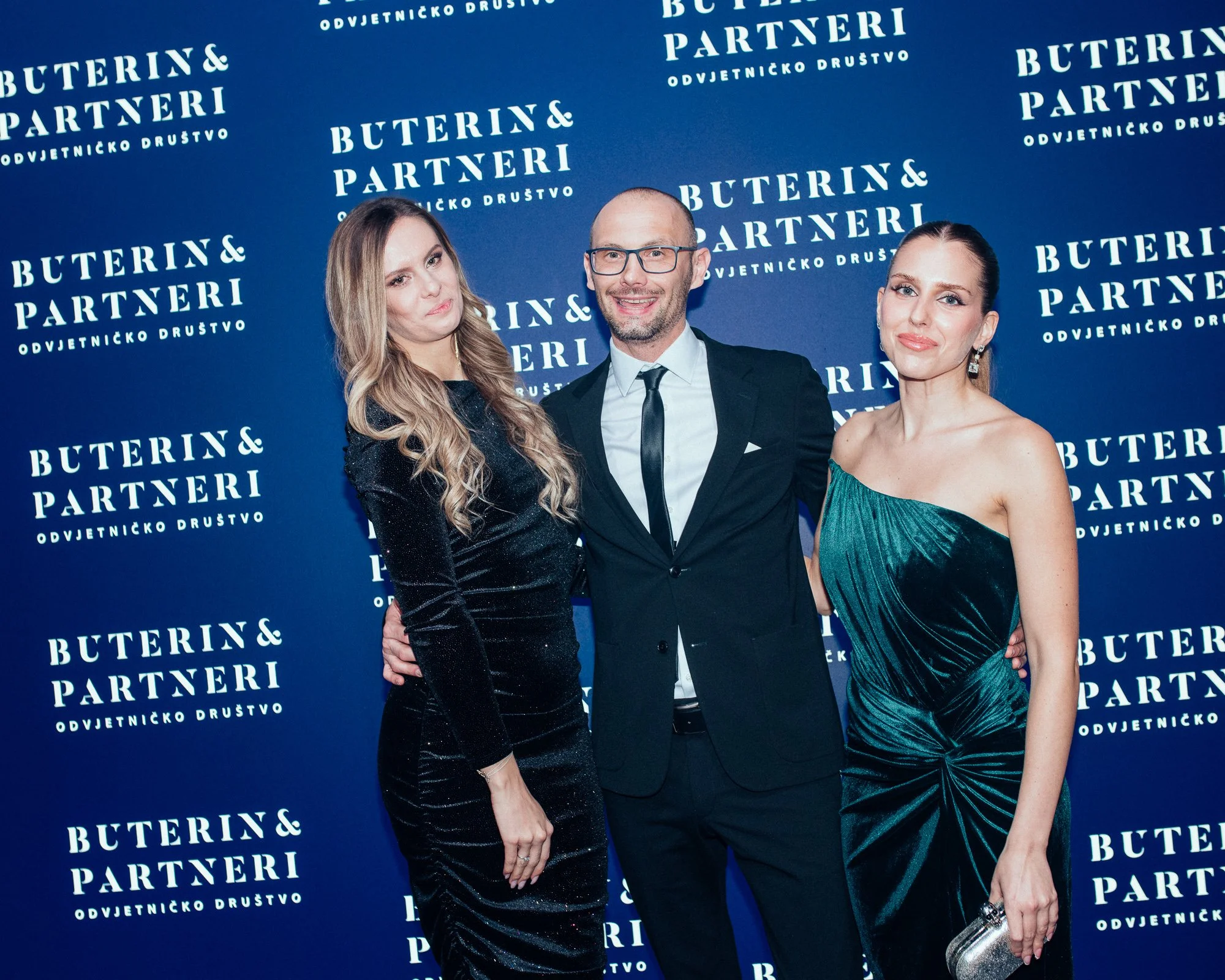 Three people, two women and one man, standing together at an event with a blue backdrop that reads 'Buterin & Partneri Odvjetničko Društvo.' The women are dressed in elegant evening gowns, and the man is in a suit. They are smiling and posing for the