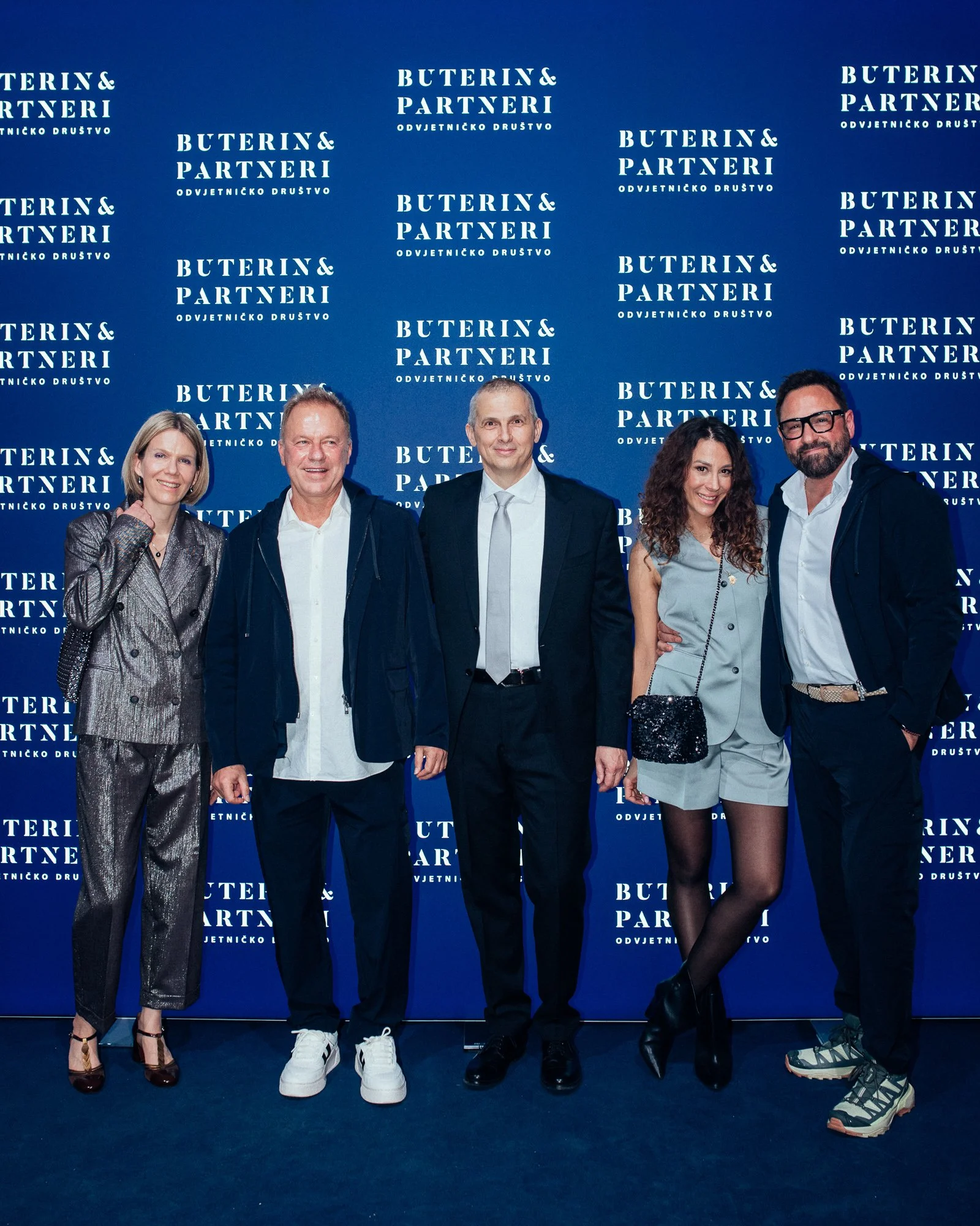 A group of five people dressed in formal and semi-formal attire standing together in front of a blue backdrop with white text that reads 'Buterin & Partneri Odvjetničko Društvo'. They are smiling and looking at the camera.