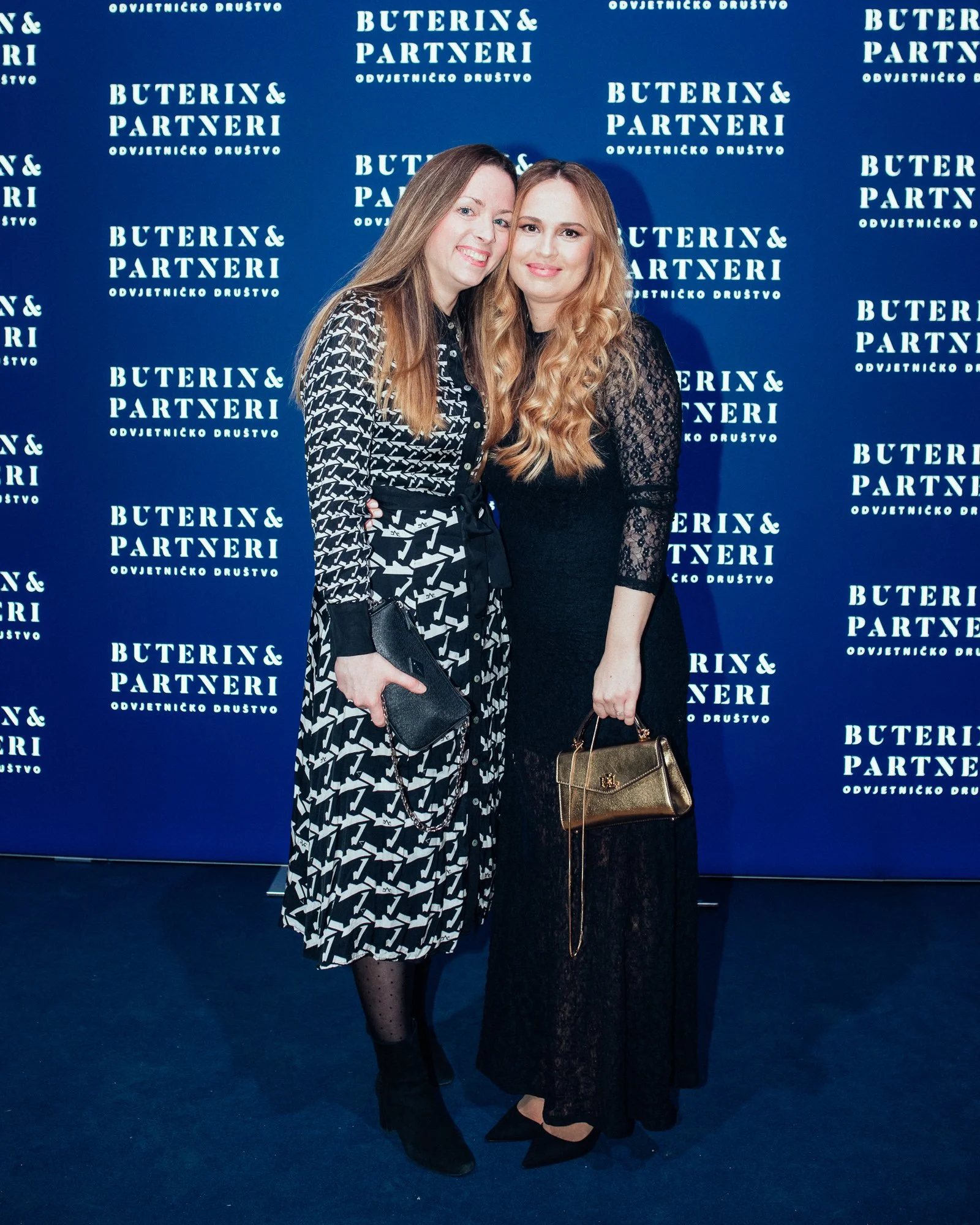 Two women standing together in front of a blue backdrop with white text, dressed in black and patterned attire, each holding a small purse.