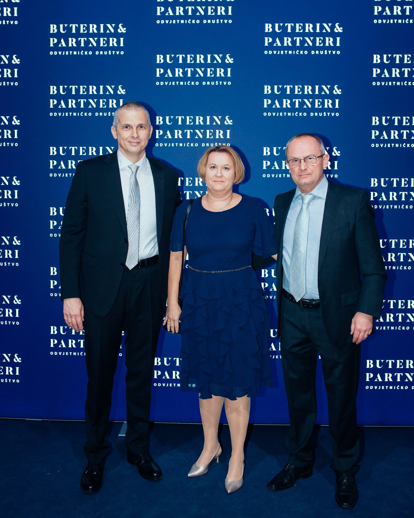 Three professionally dressed individuals, two men and one woman, standing in front of a blue backdrop with the words 'BUTEIN & PARTNERI' repeated multiple times.