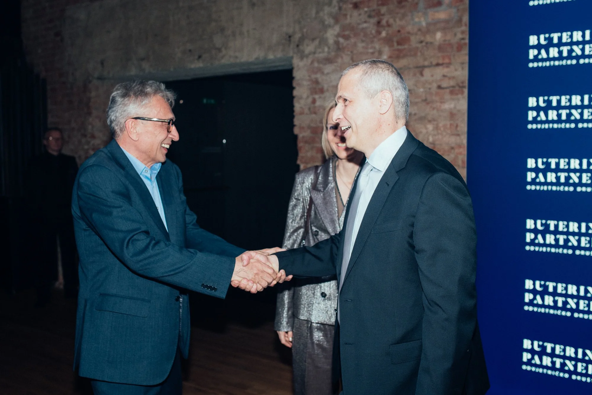 Two men in suits shaking hands and smiling at each other at a professional event, with a woman standing behind them and a panel or screen with the text 'Butterfly Partners' in the background.