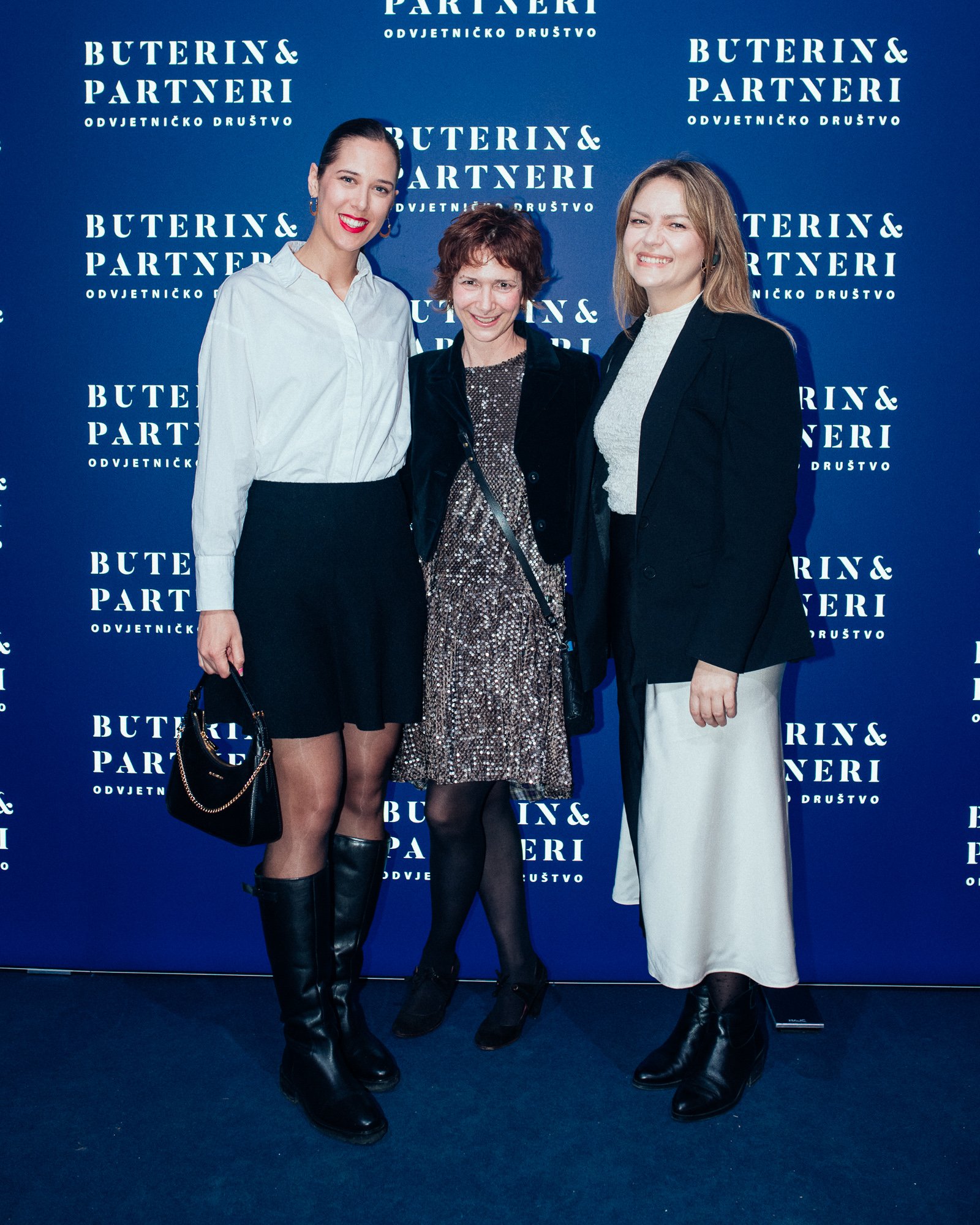 Three women standing together in front of a backdrop with the text 'Buterin & Partneri'. They are smiling, dressed in semi-formal attire, and one is holding a handbag.