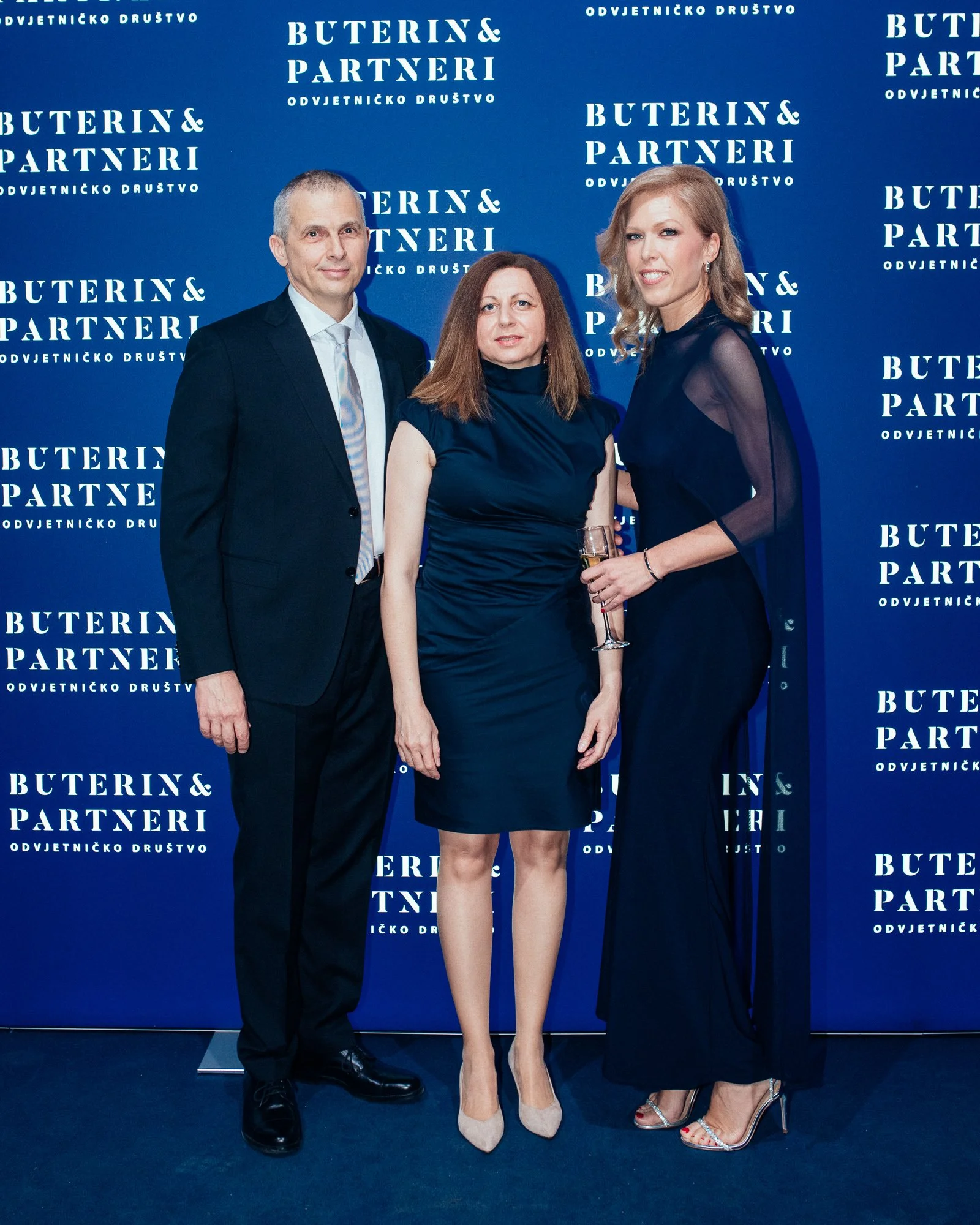 Three formally dressed people standing in front of a blue backdrop with white text that reads "Buterin & Partneri." The man on the left is wearing a black suit and tie, the woman in the middle is wearing a fitted dark blue dress, and the woman on the