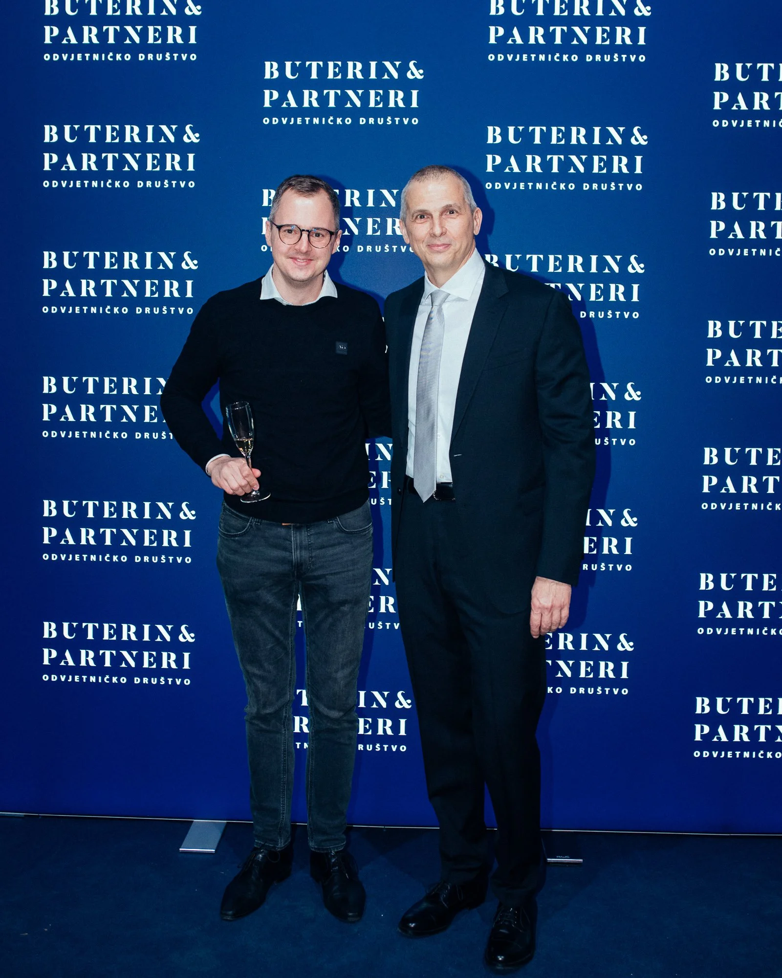 Two men at a formal event standing in front of a blue backdrop with white text that reads 'BUTERIN & PARTNERI' and 'ODVJETNIČKO DRUŠTVO.' One man is wearing glasses, a black sweater, jeans, and holding a glass of champagne. The other man is dressed i