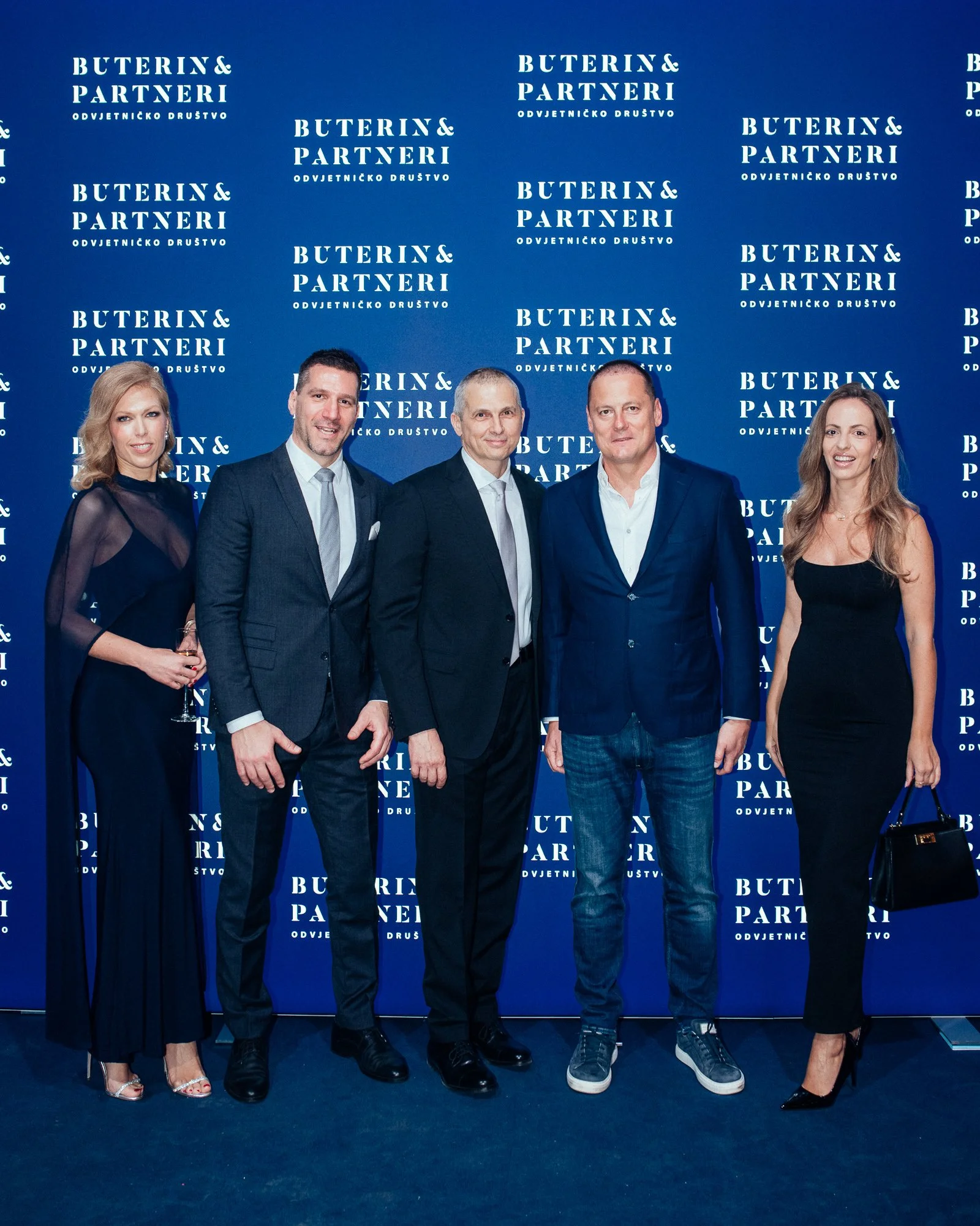 Group of five people dressed in formal and semi-formal attire standing in front of a blue backdrop with the logo 'BUTERIN & PARTNERI' repeatedly printed on it.