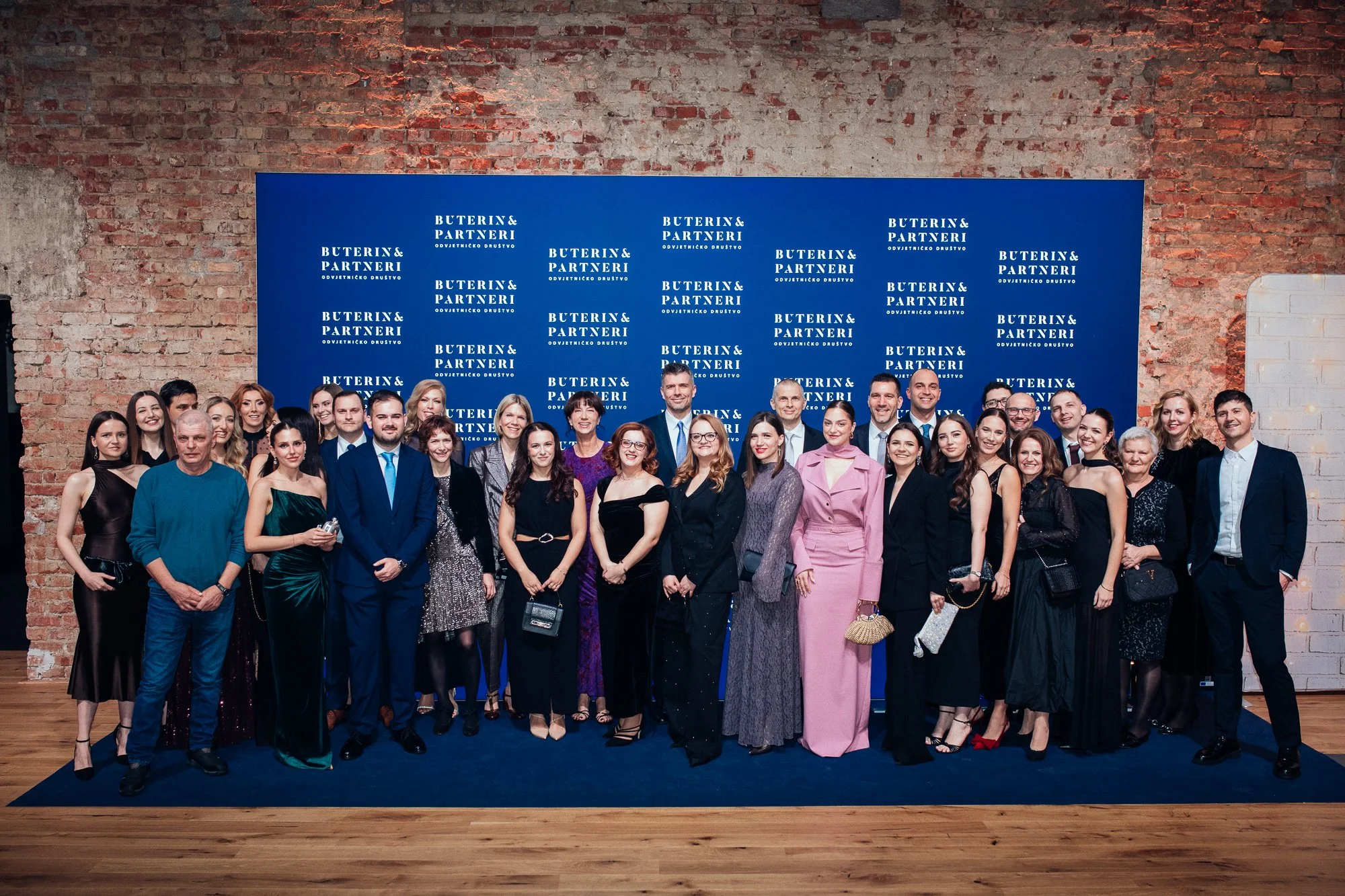 Group of people dressed in formal attire posing for a photo at an event with a blue backdrop that reads 'Buterin & Partneri' against a brick wall.