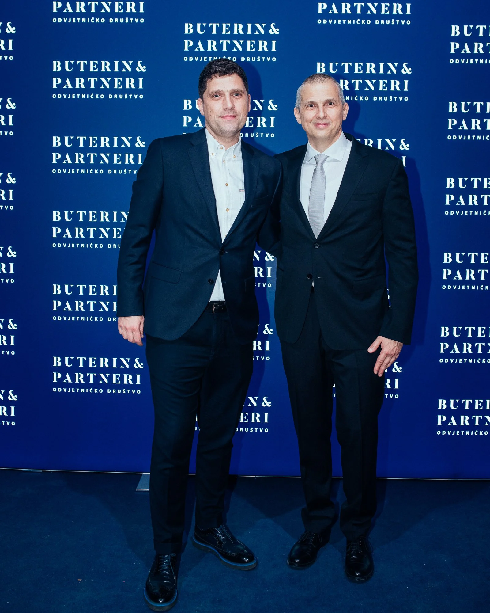 Two men in dark suits standing in front of a blue background with white text that reads 'Buterin & Partneri ODVJETNICKO DRUŠTVO'.