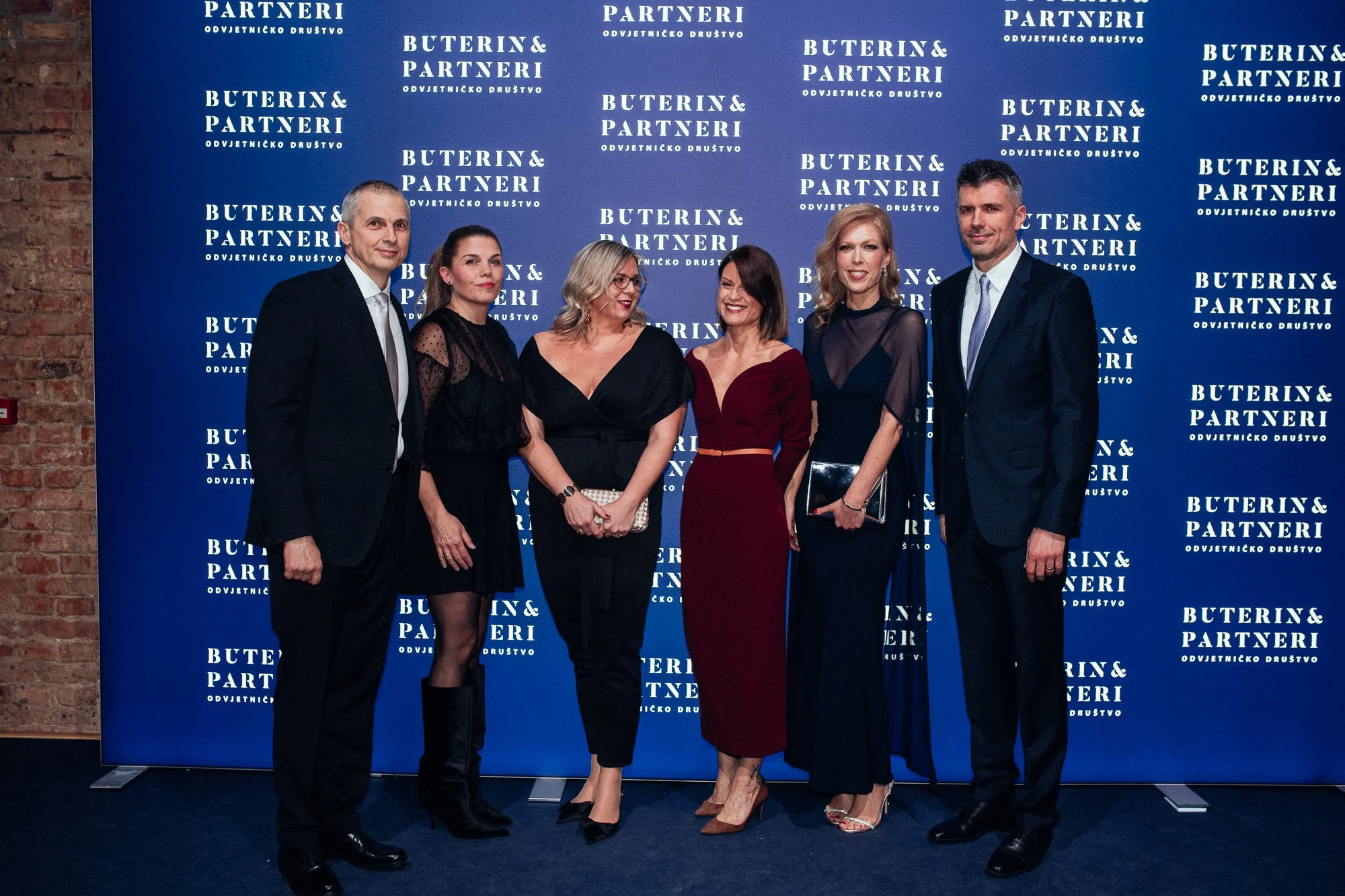 Group of six professionally dressed people standing together at an event with a blue backdrop featuring the names 'Buterin & Partneri' repeated multiple times.