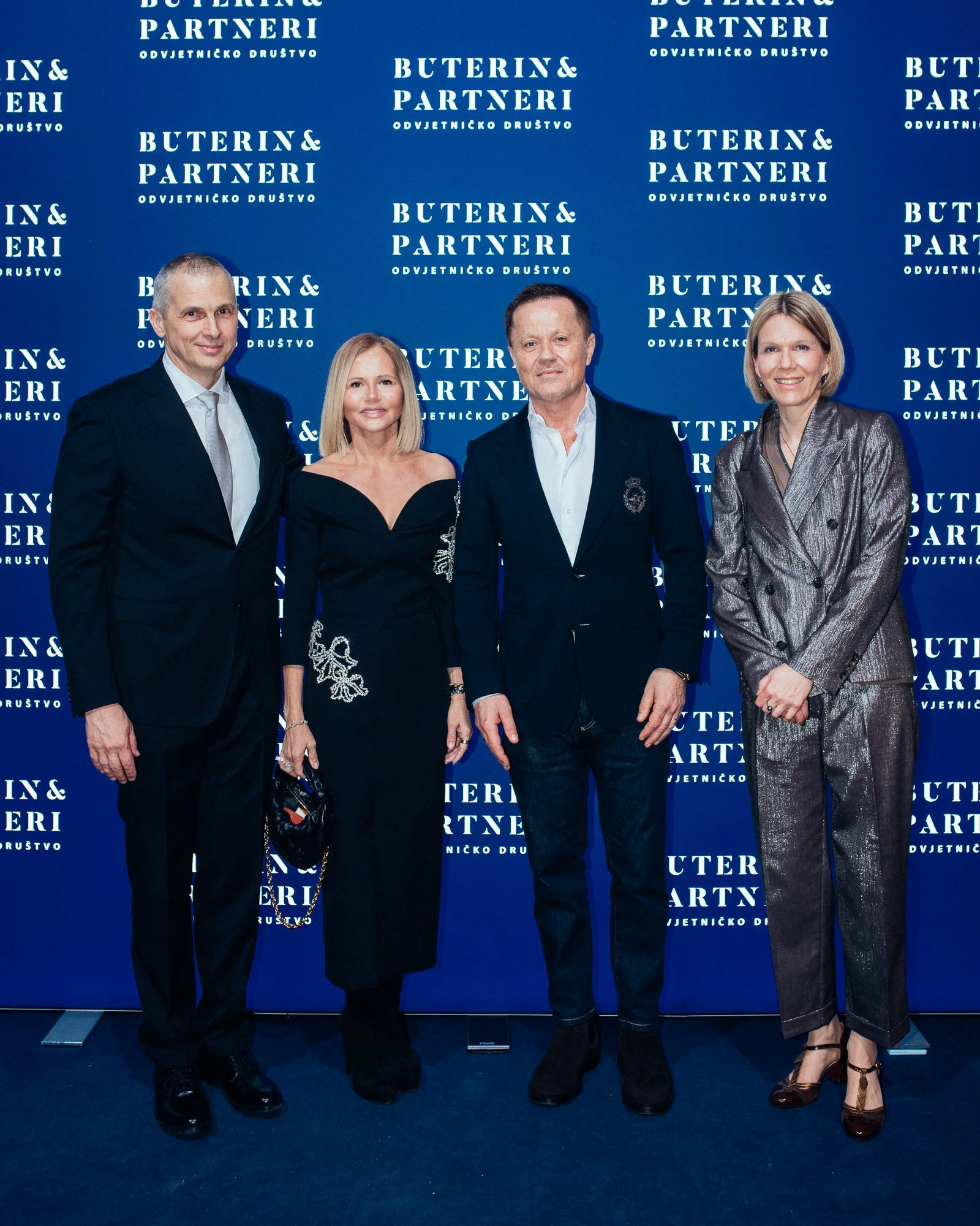 Four formally dressed individuals standing in front of a blue backdrop with the logo and text "Buterin & Partneri" repeatedly printed on it.