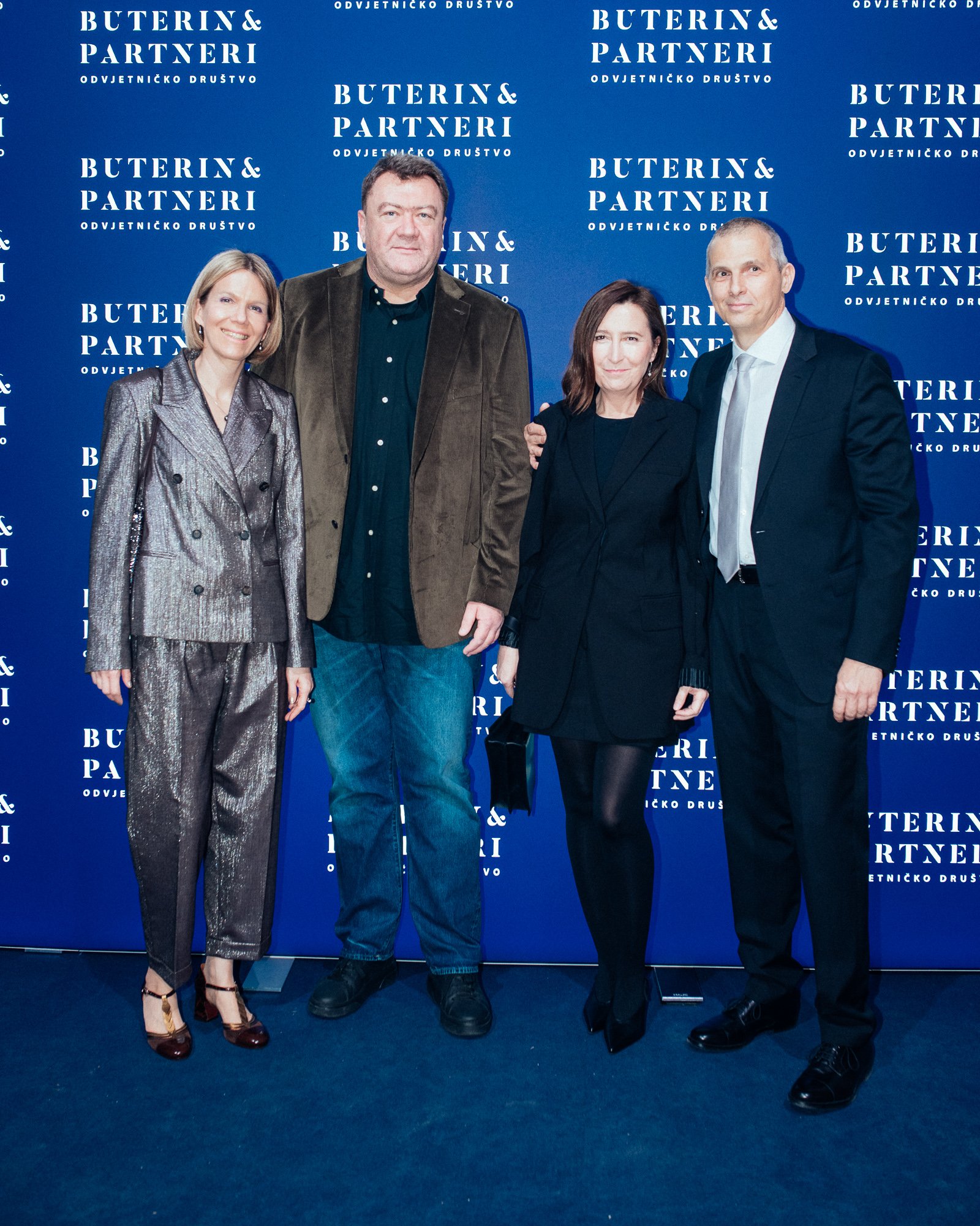 Four suited individuals standing together at an event with a blue backdrop featuring the text 'Buterin & Partneri' repeated multiple times. They are smiling, with arm links, and wearing formal attire.